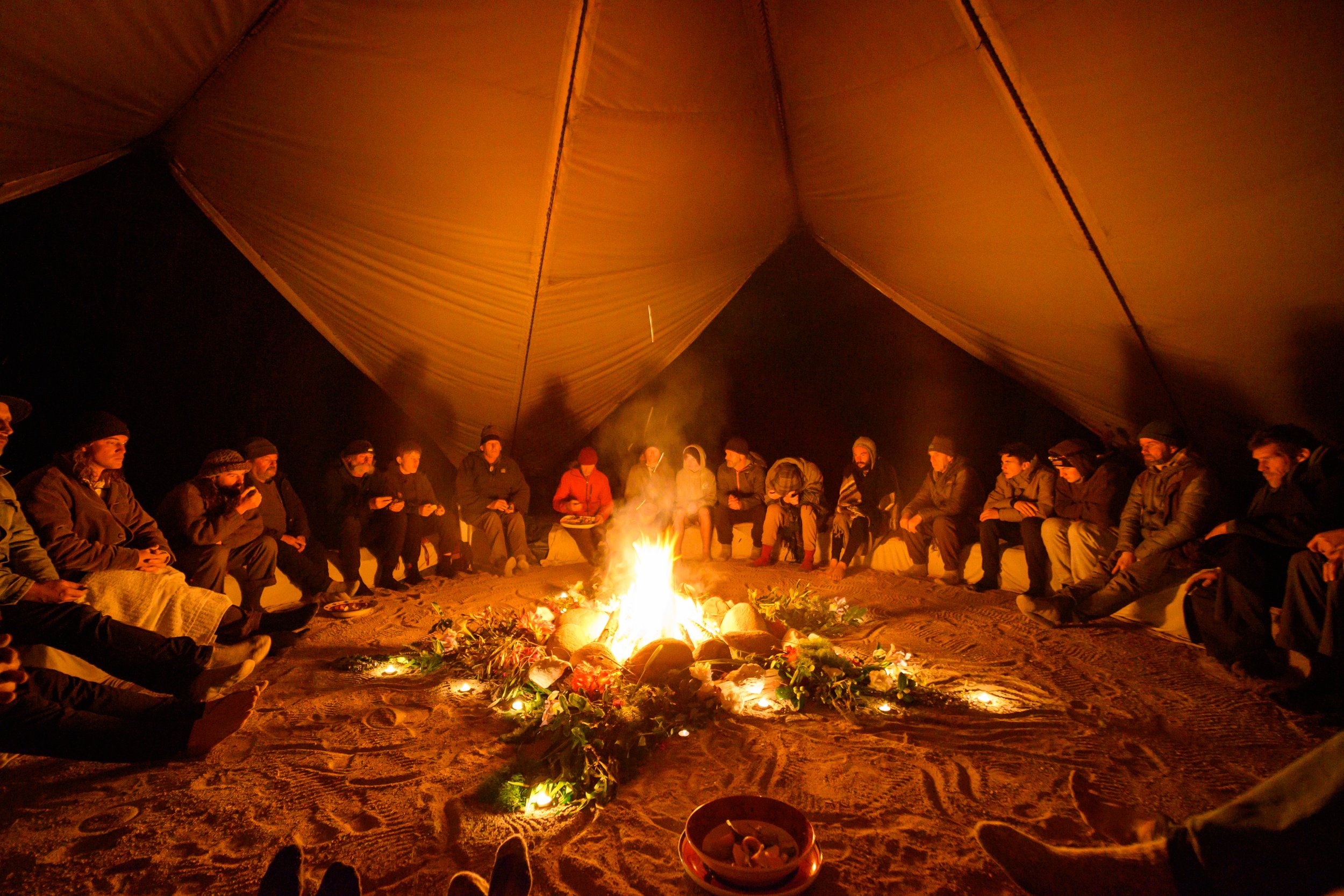 People sitting in a circle around a campfire inside a large tent at night during a Wilderness Rites of Passage event in New Zealand