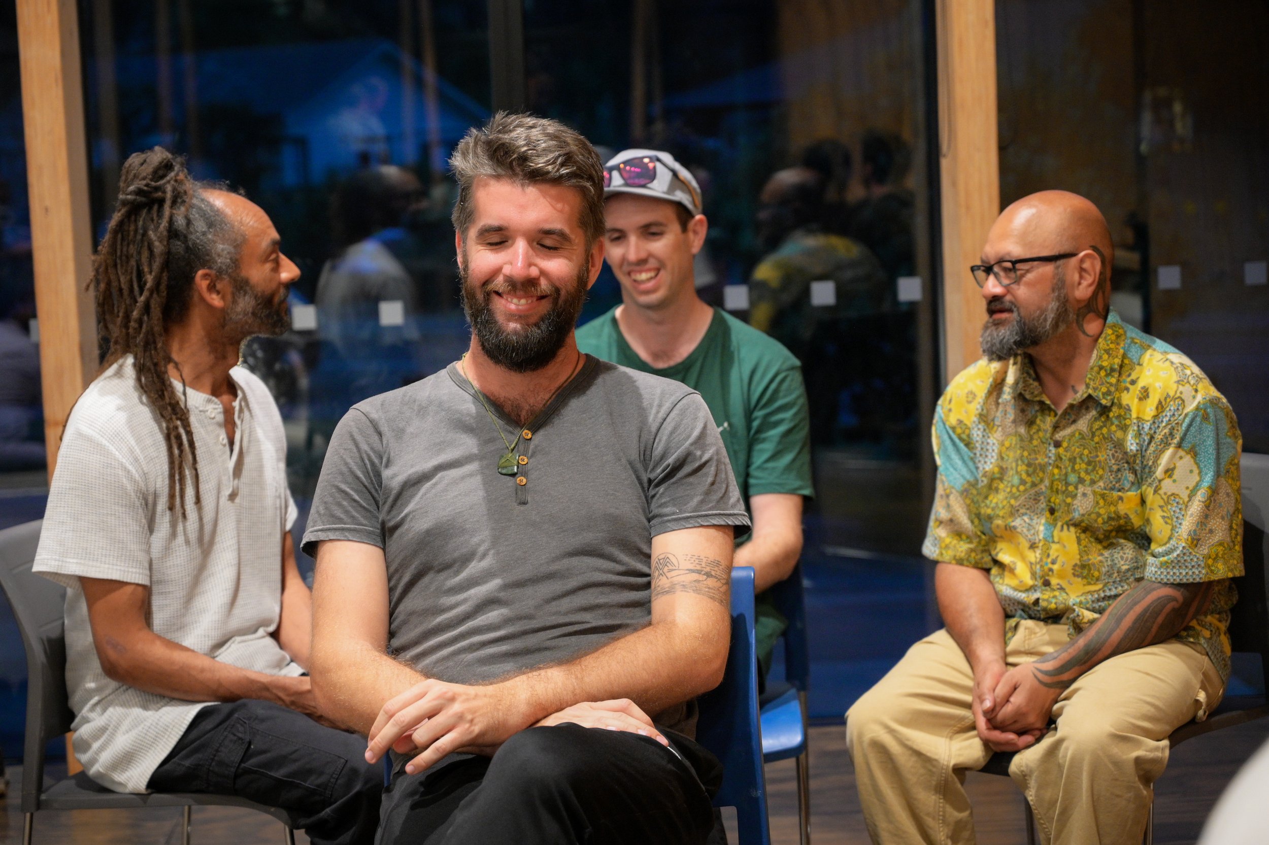 Four men sitting in a circle, smiling and engaging in conversation at an indoor gathering or event.