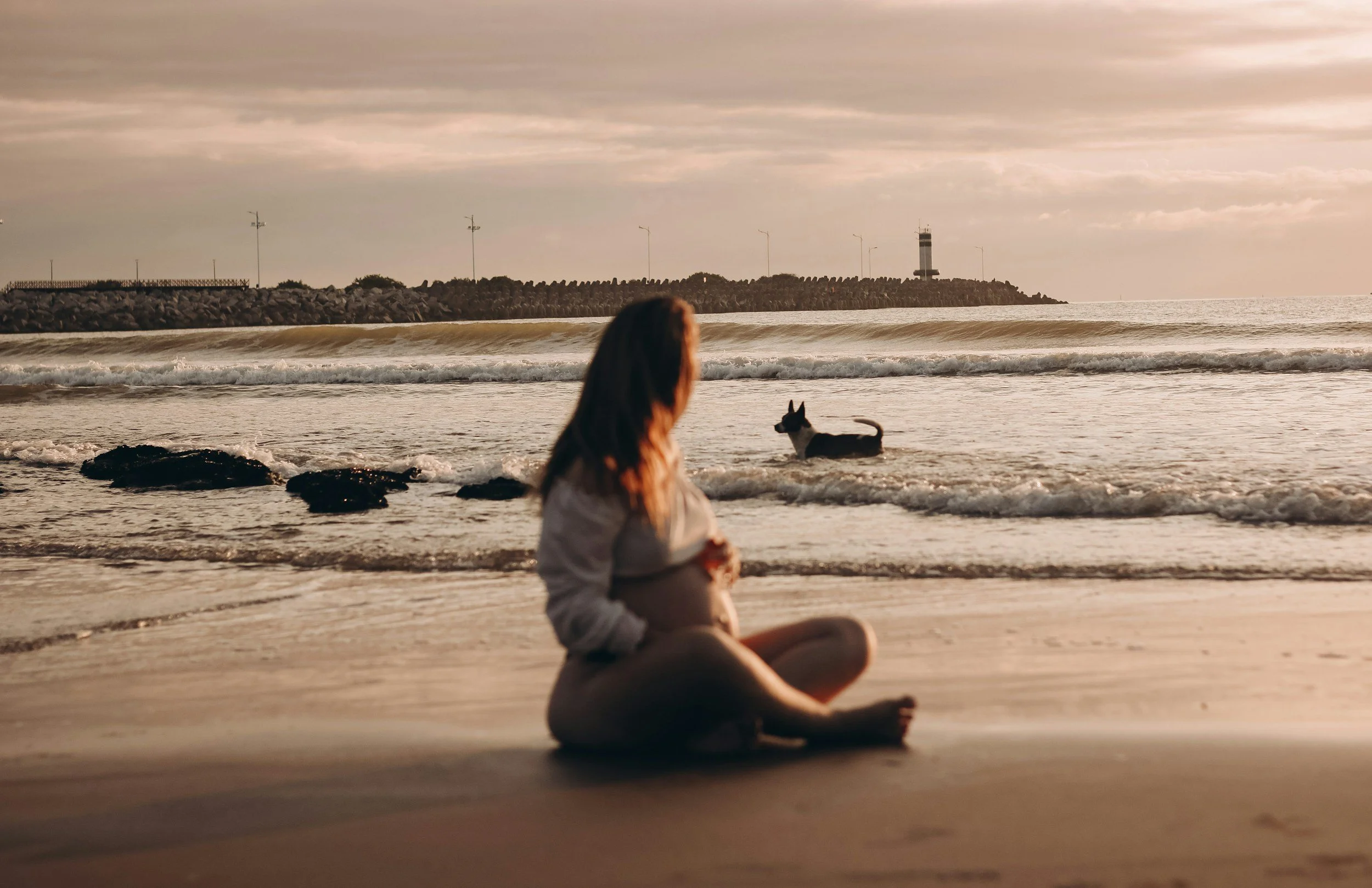 A pregnant woman sitting on the sandy beach looking at the ocean, with a dog in the water nearby and a lighthouse in the distance during sunset.