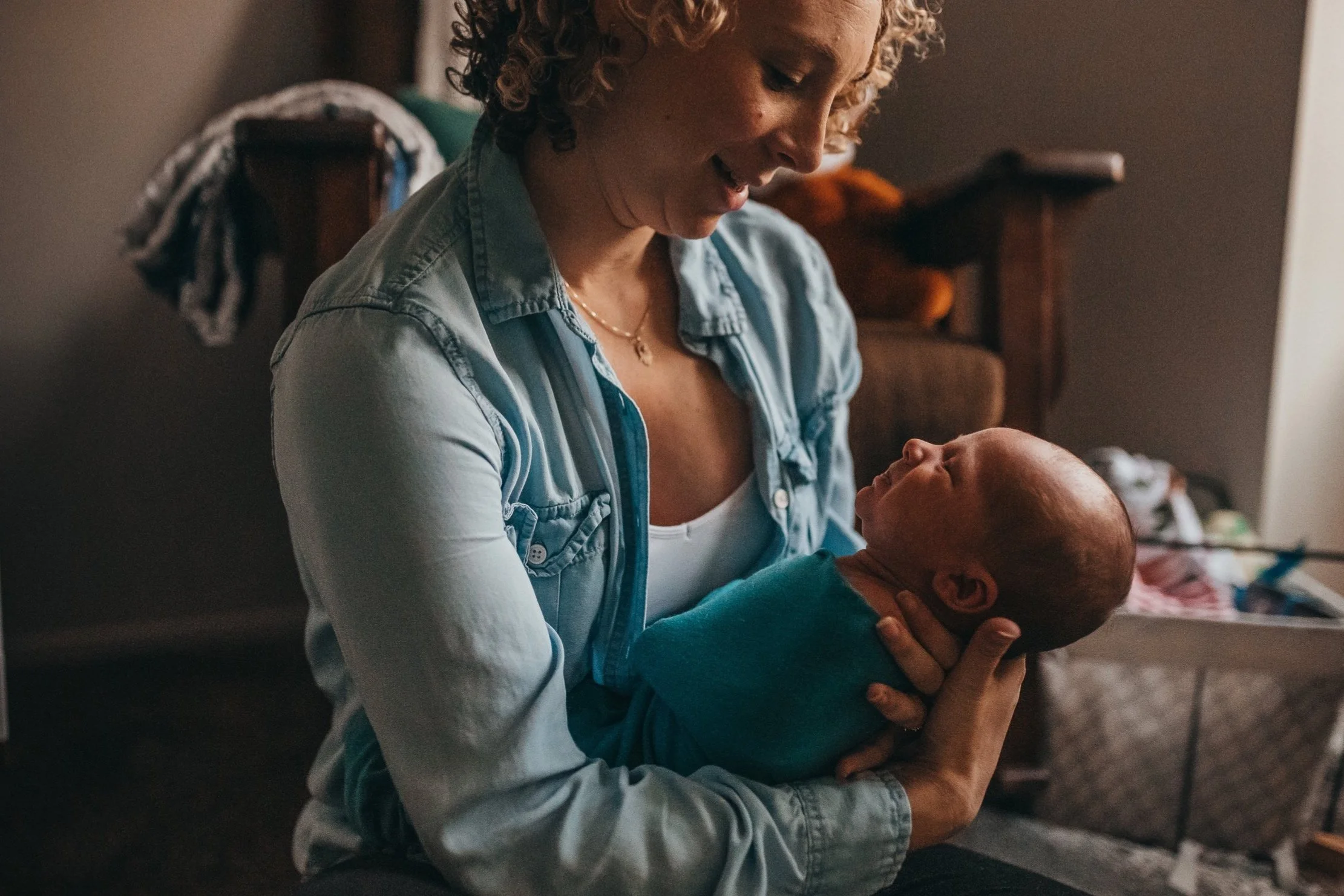 A woman with curly blonde hair smiling at a baby she is holding in her lap, with a dark background and a piece of furniture in the background.