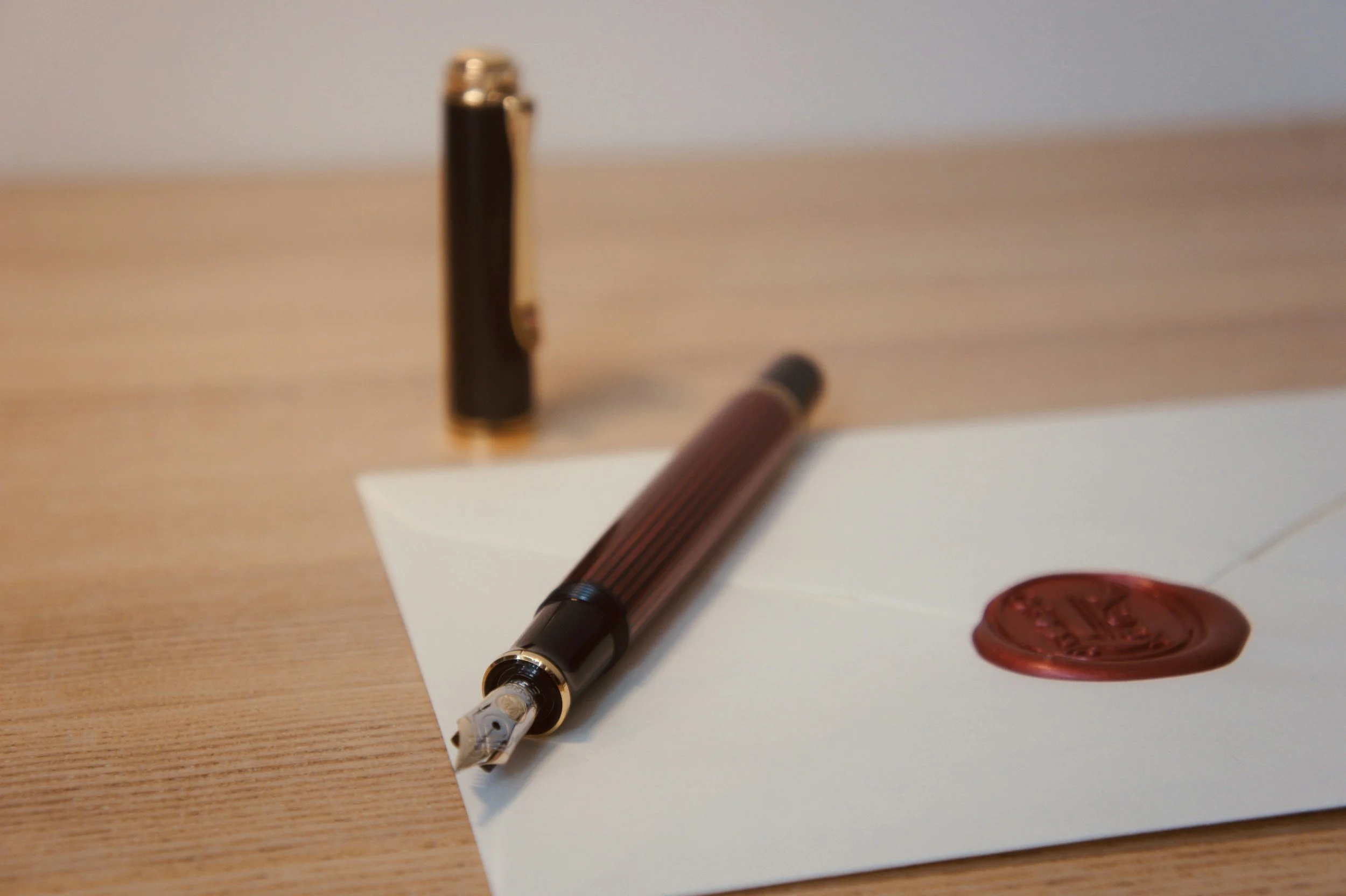 A fountain pen with a silver and black design resting on a white sheet of paper, a red wax seal, and a wax seal stamp on a wooden surface.