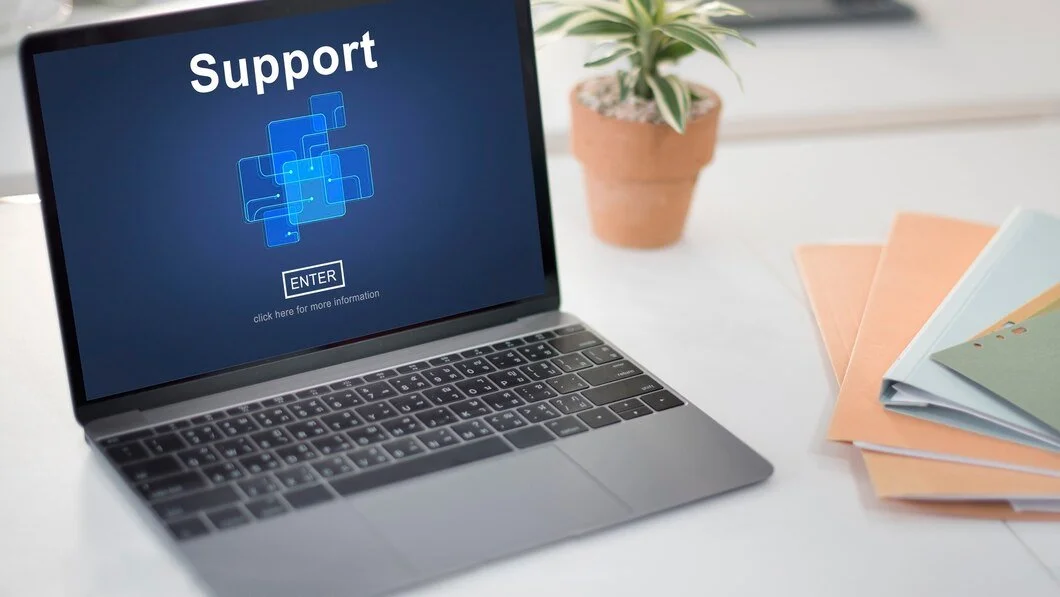 Laptop with a support page on the screen, next to a potted plant and some colorful folders on a white desk.