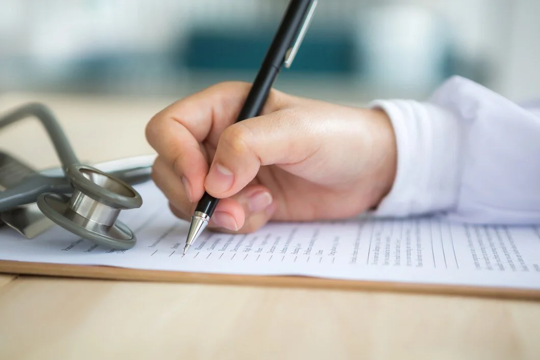 A person in a white medical coat is writing on a form with a black pen. There is a stethoscope on the clipboard beside the paper.