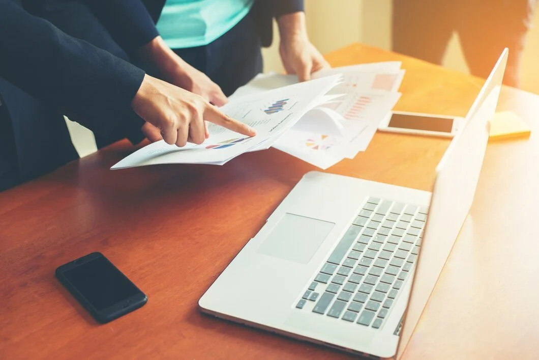 People reviewing financial documents with a laptop, smartphone, and tablet on a wooden table.