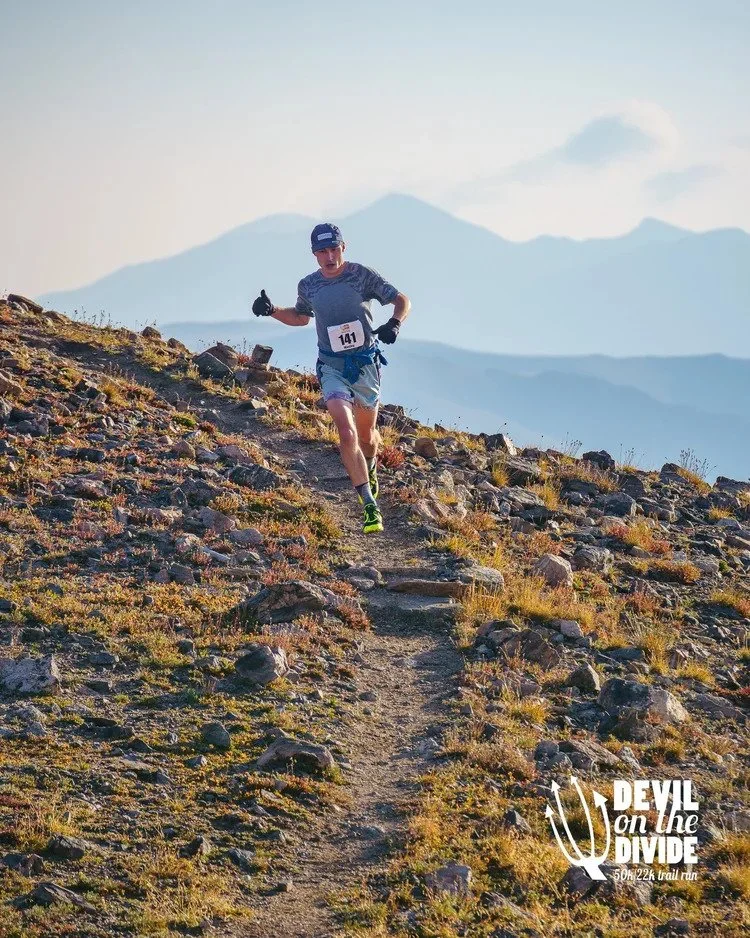 A male trail runner wearing a blue cap, gray shirt, and shorts running uphill on a rocky trail with mountains in the background during daylight. There is a logo in the bottom right corner for 'Devil on the Divide' 50k/22k trail run.