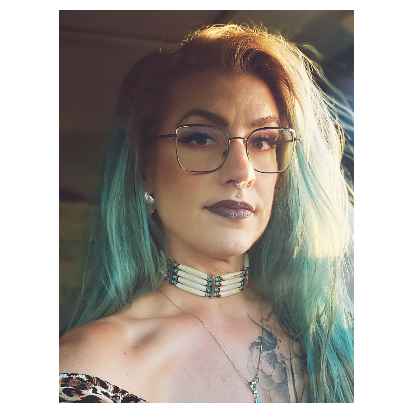 A woman with light green and blue hair wearing glasses, dark lipstick, a choker necklace, tattoos, and a leopard-print top, looking at the camera.
