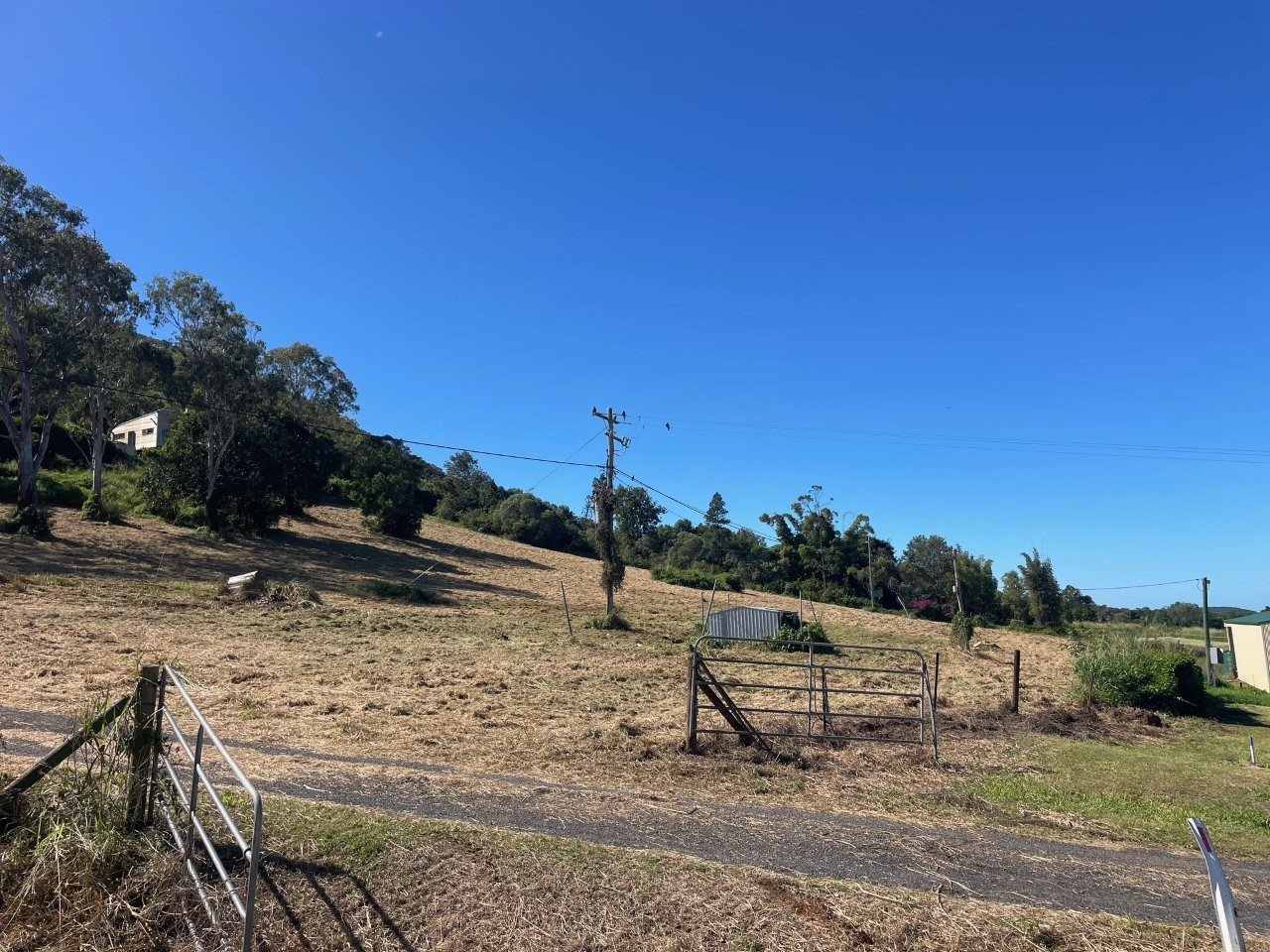 A hilly landscape with a clear blue sky, trees, a utility pole, a small building, a metal gate, and a fence on a rural property.