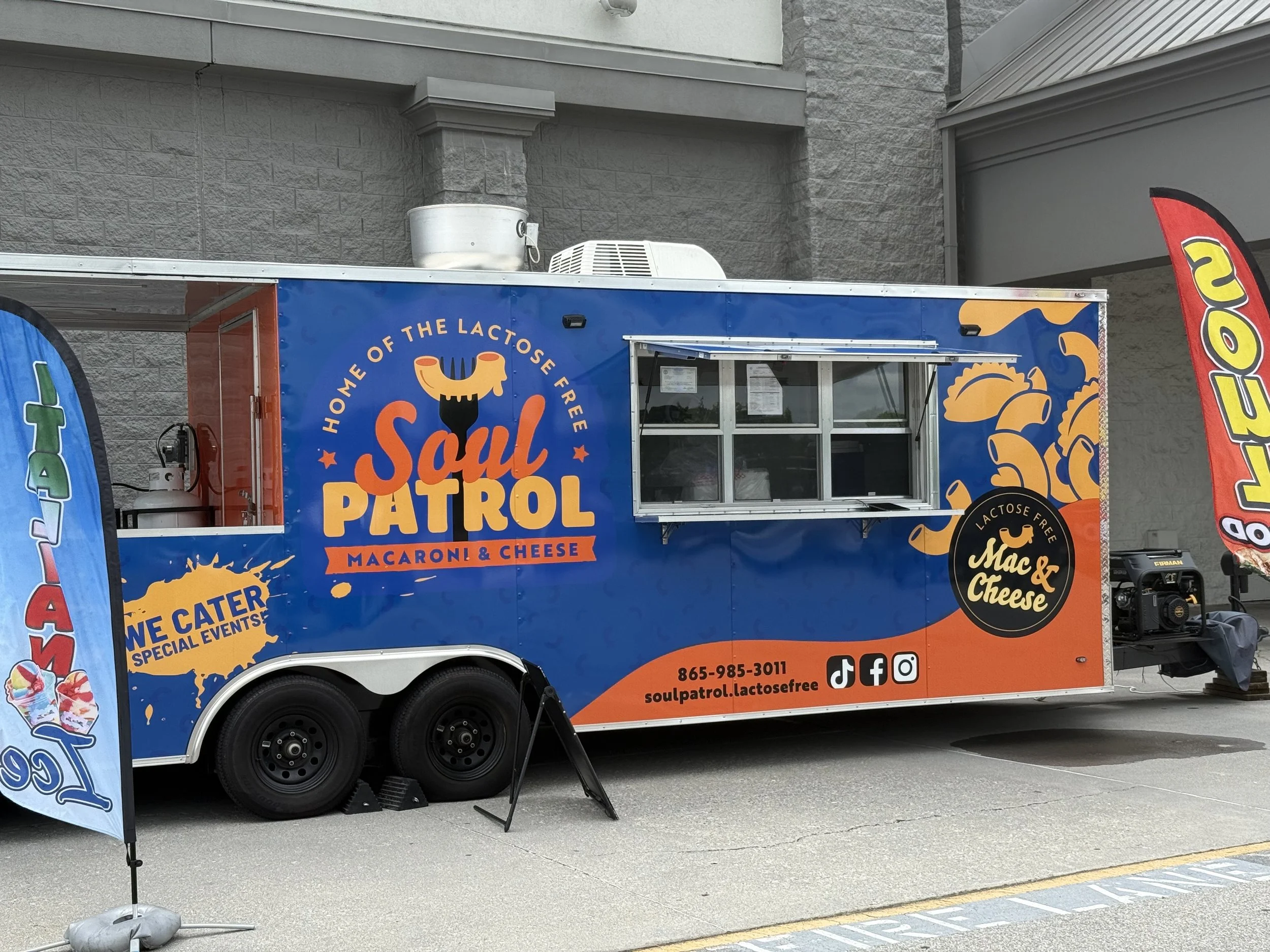 Bold, bright, and vibrant orange and blue, Soul Patrol food truck serving lactose-free macaroni and cheese, parked on the street with flags on each side advertising the food and special events.