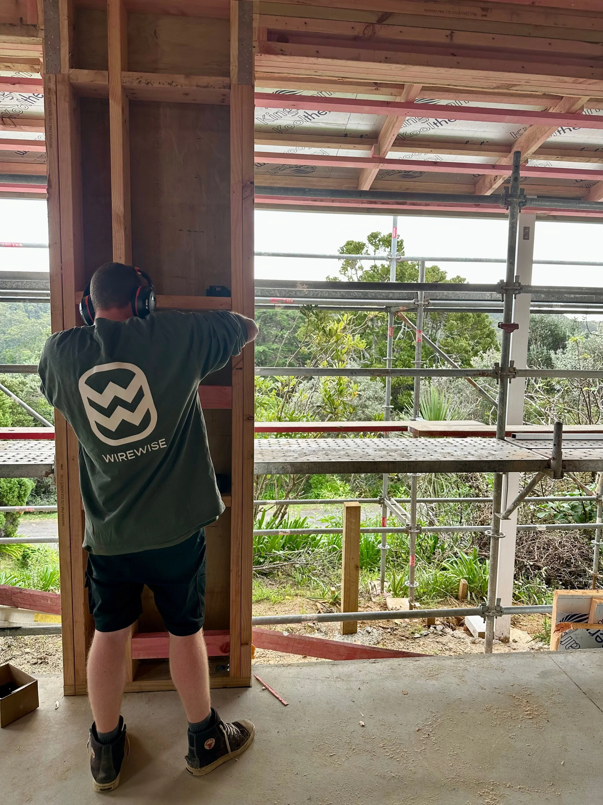 A person with a dark shirt and shorts wearing boots and headphones working on framing a wall inside a house under construction. The room has exposed wooden beams, scaffolding, and a view of trees outside.