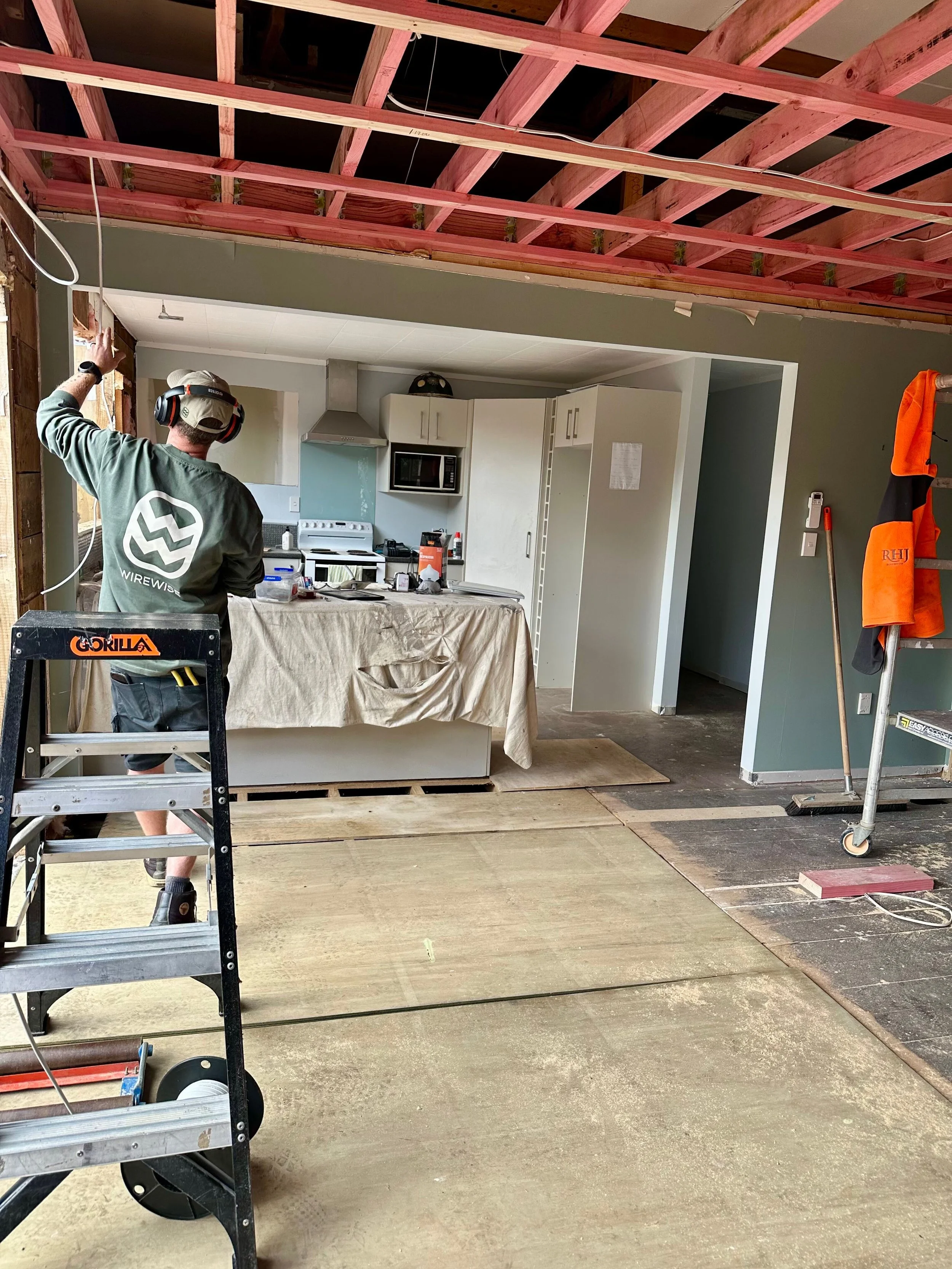 Construction worker in a green shirt and cap working in a partially renovated kitchen with exposed wooden ceiling framework, white cabinets, and a countertop covered with a protective cloth.