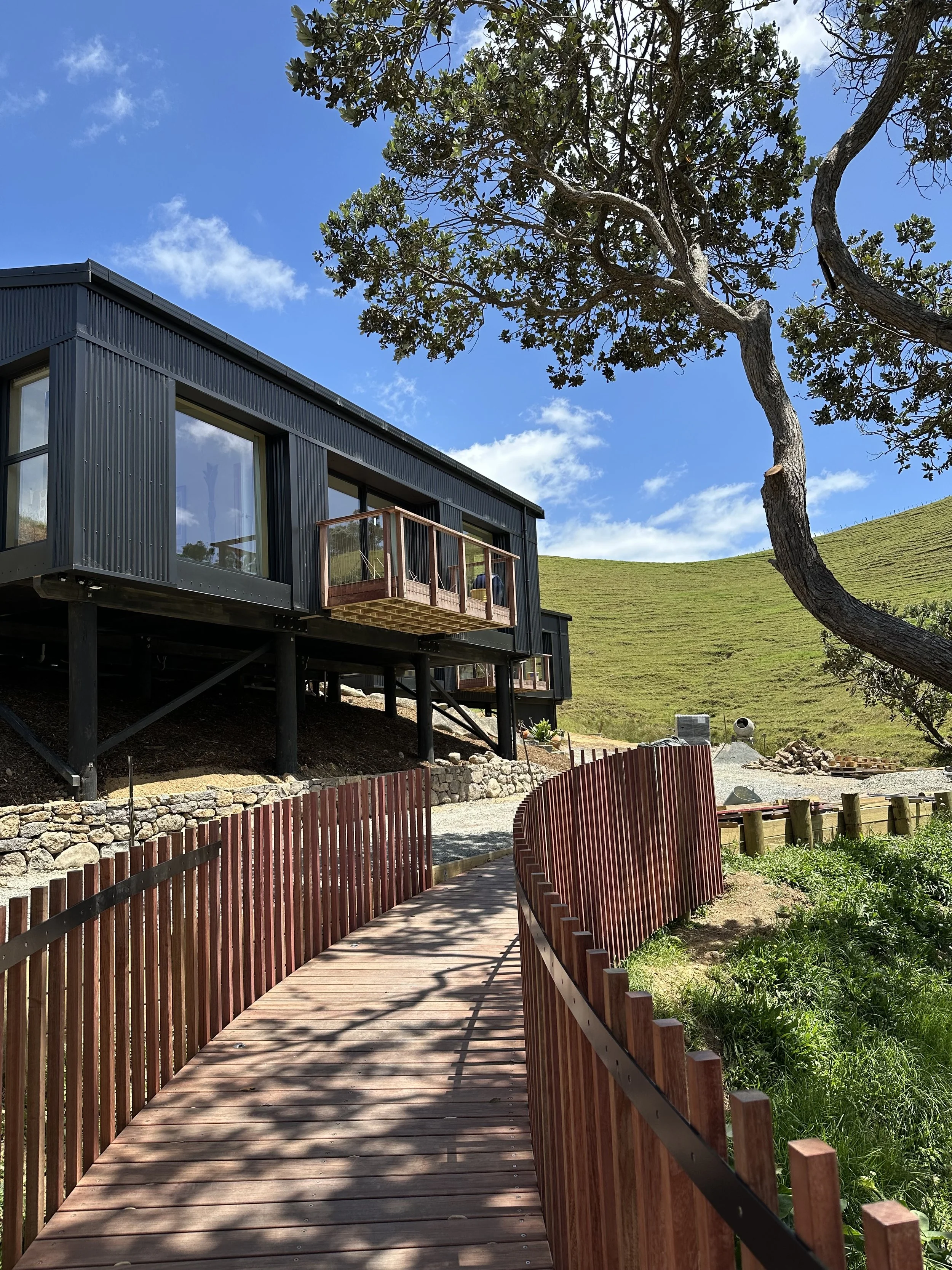 Wooden walkway with red railings leading to a modern black house elevated on stilts, surrounded by green hills, a large tree, and a blue sky with some clouds.