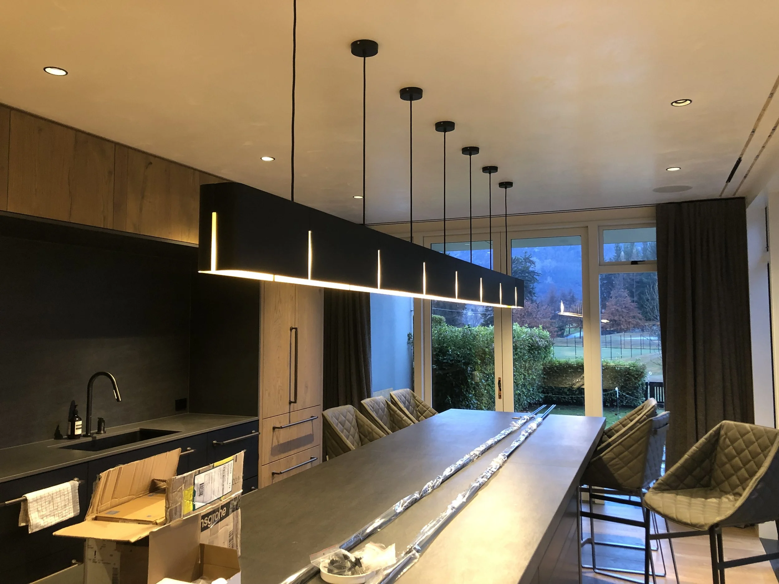 Modern dining room with a long island table, cushioned chairs, black and wood cabinetry, a unique black hanging light fixture, and large windows showing an outdoor landscape with trees and a field.