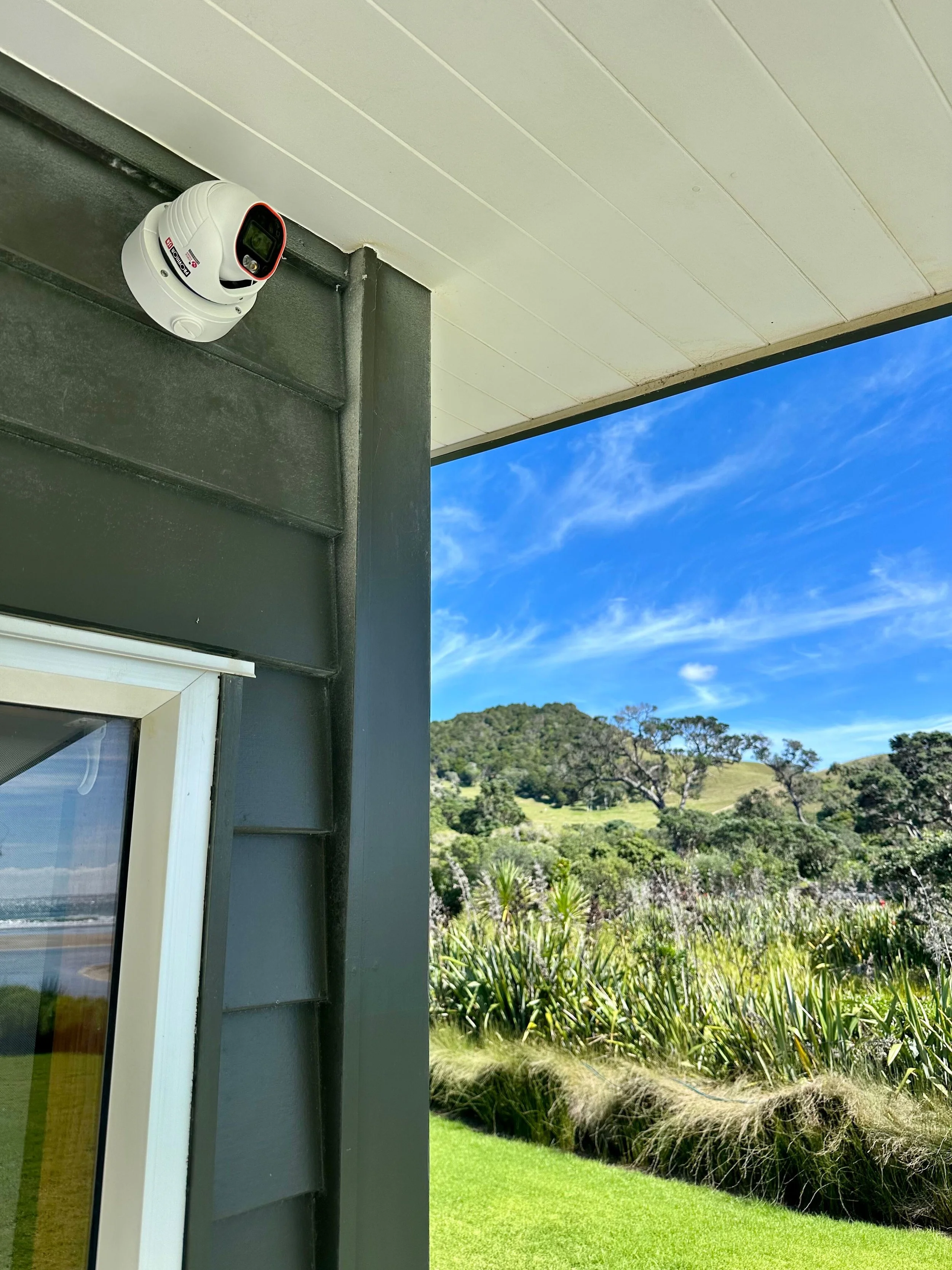 A security camera mounted on the side of a building, with a view of green hills, trees, and a blue sky with scattered clouds in the background.