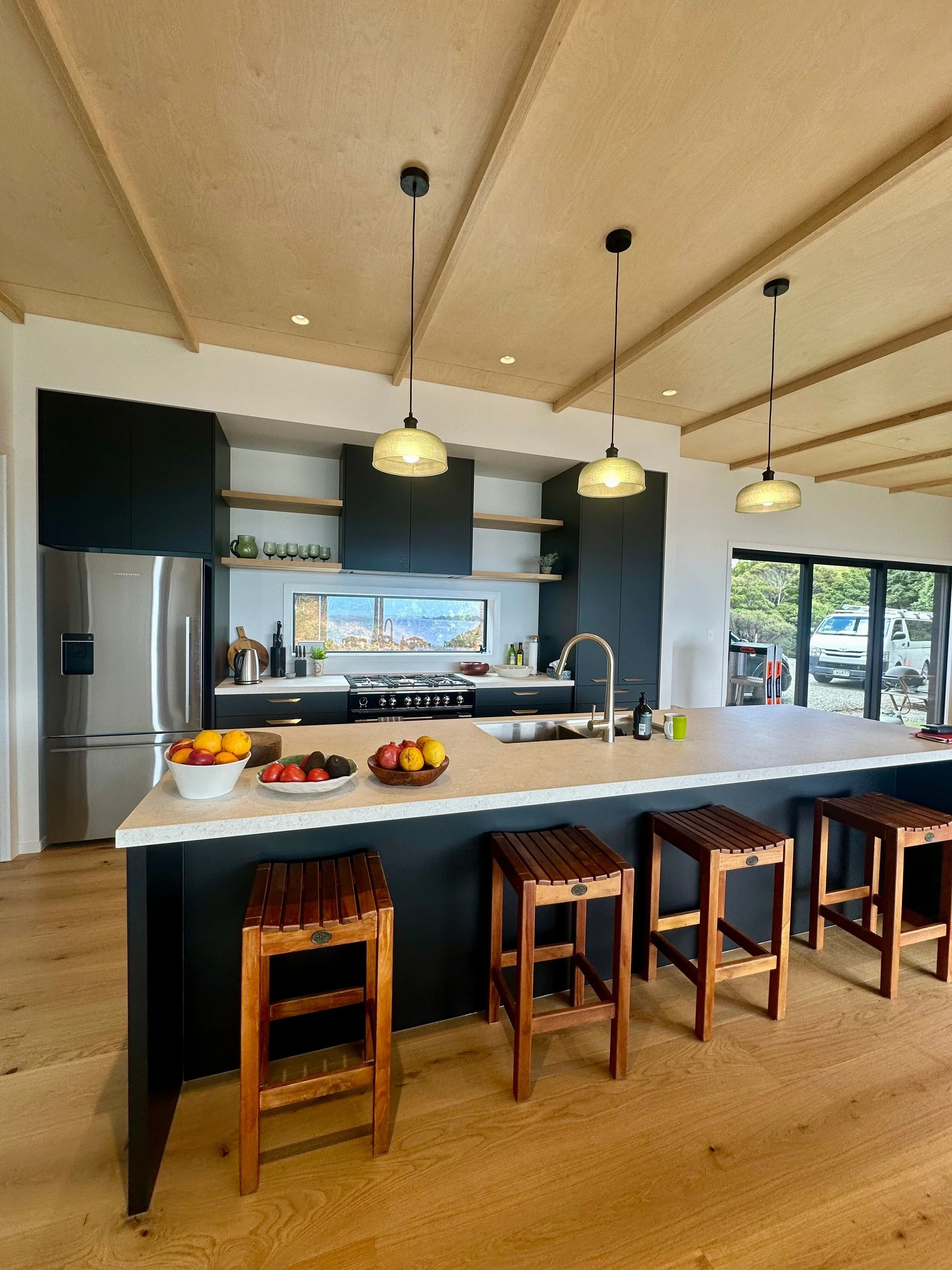 Modern kitchen with a large island, black cabinetry, and hanging pendant lights. The island has bowls of fruit and a wooden stool seating area. Large windows and sliding glass door reveal outdoor greenery and parked cars.