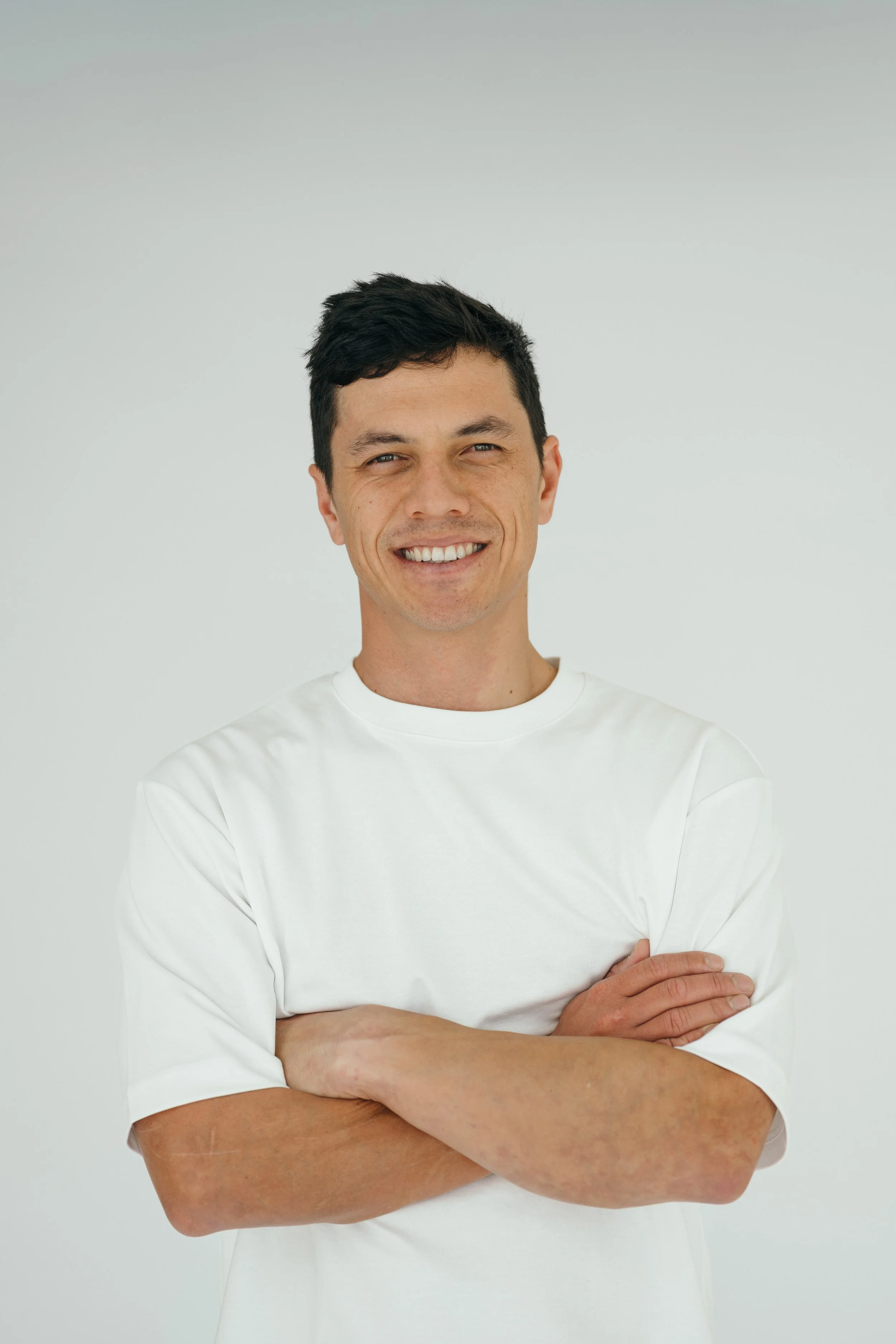 A man with short dark hair and light skin, smiling, wearing a white long-sleeve shirt with arms crossed, against a plain light gray background.