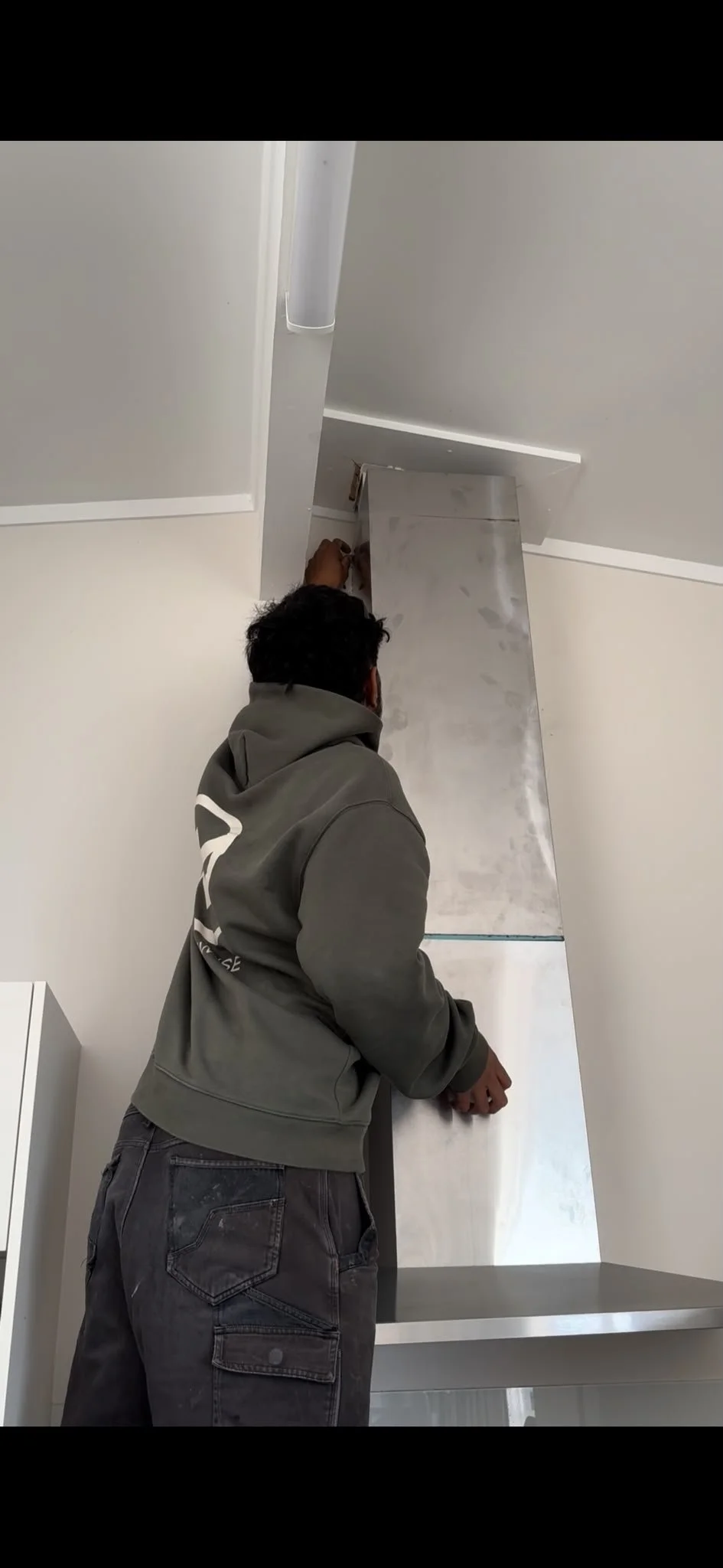 A person in a gray hoodie working on a kitchen cabinet installation with stainless steel backsplash and white walls.