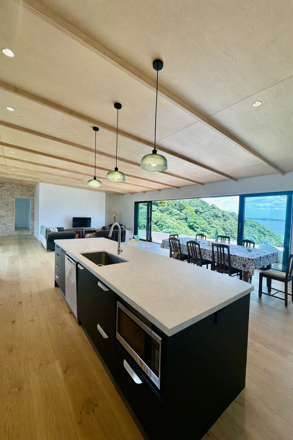 Open-concept living space with a kitchen island, dining area, and living room, large windows showing a scenic view of hills and water, with wooden ceiling and floor.