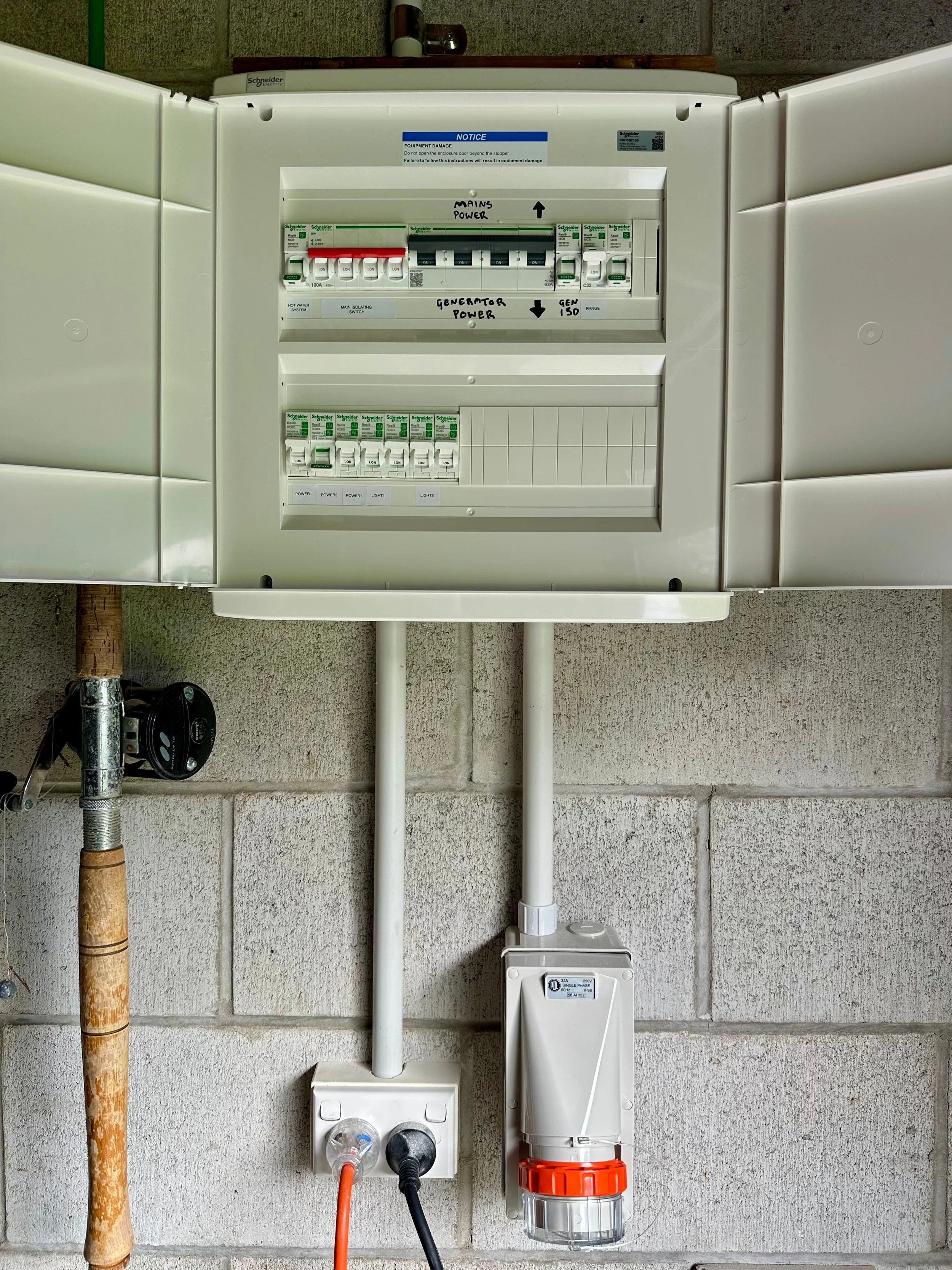 Electrical panel with various switches and labels, mounted on a concrete wall, with wires and a pipe below.