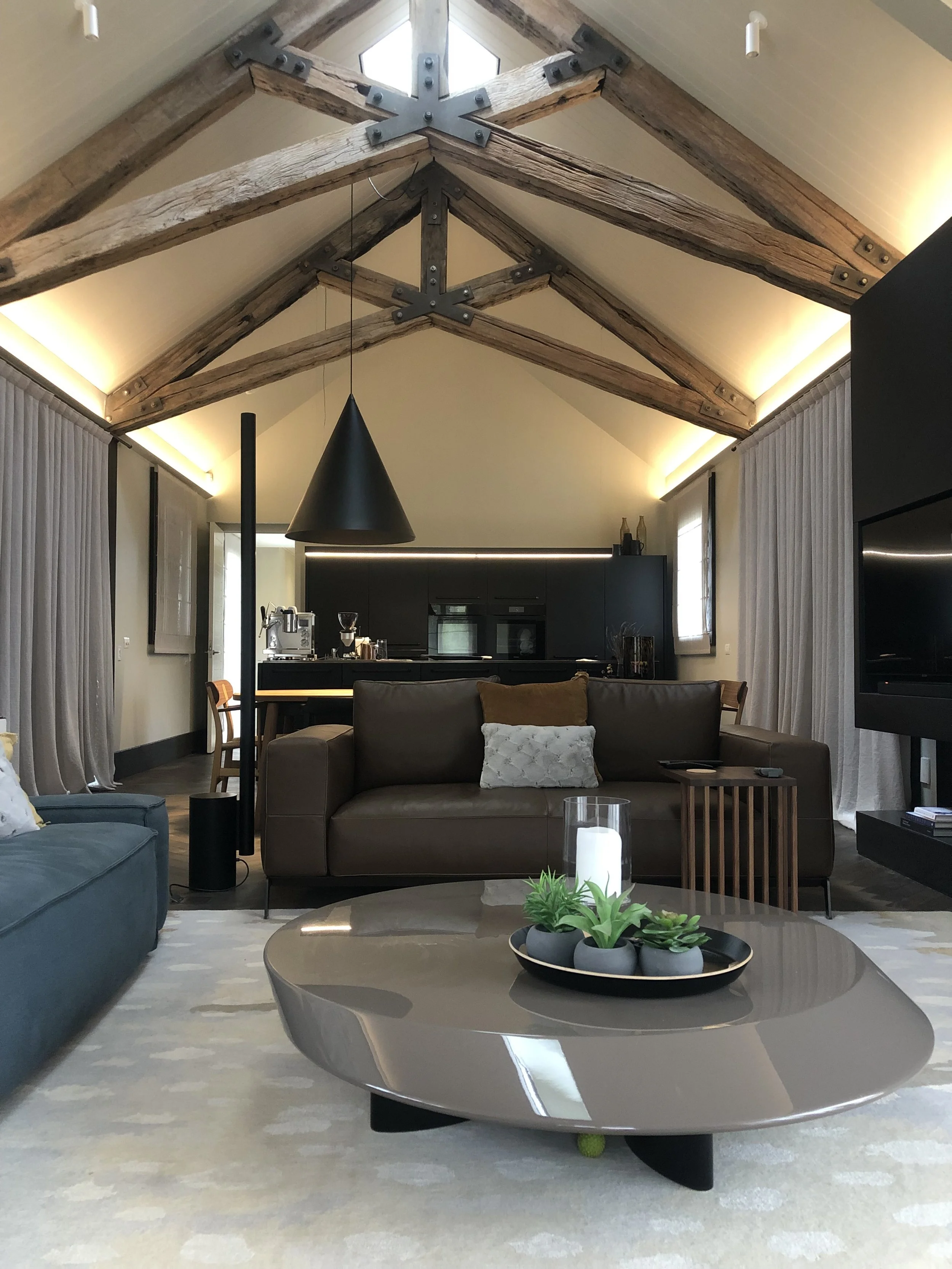 Living room with a vaulted ceiling featuring exposed wooden beams, a brown sofa with decorative pillows, a modern black coffee table with a tray of succulents and a candle, and a black pendant light hanging from the ceiling. Curtains cover windows on either side of the room.