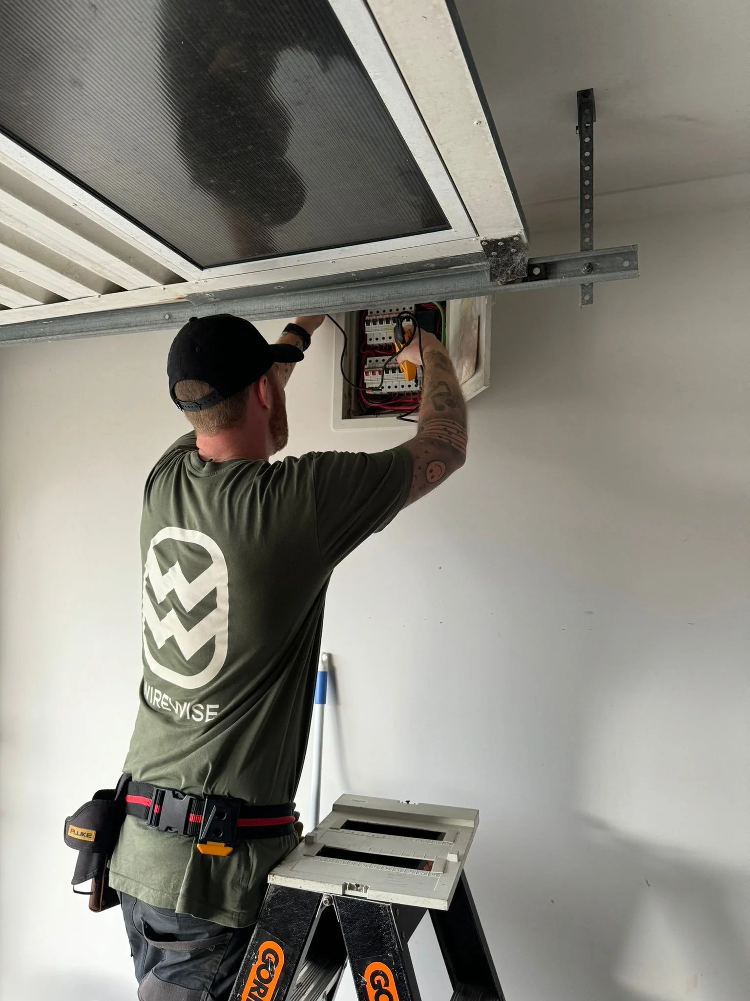 A technician working on an electrical panel or control box mounted on a wall, standing on a ladder, with a multimeter in hand.