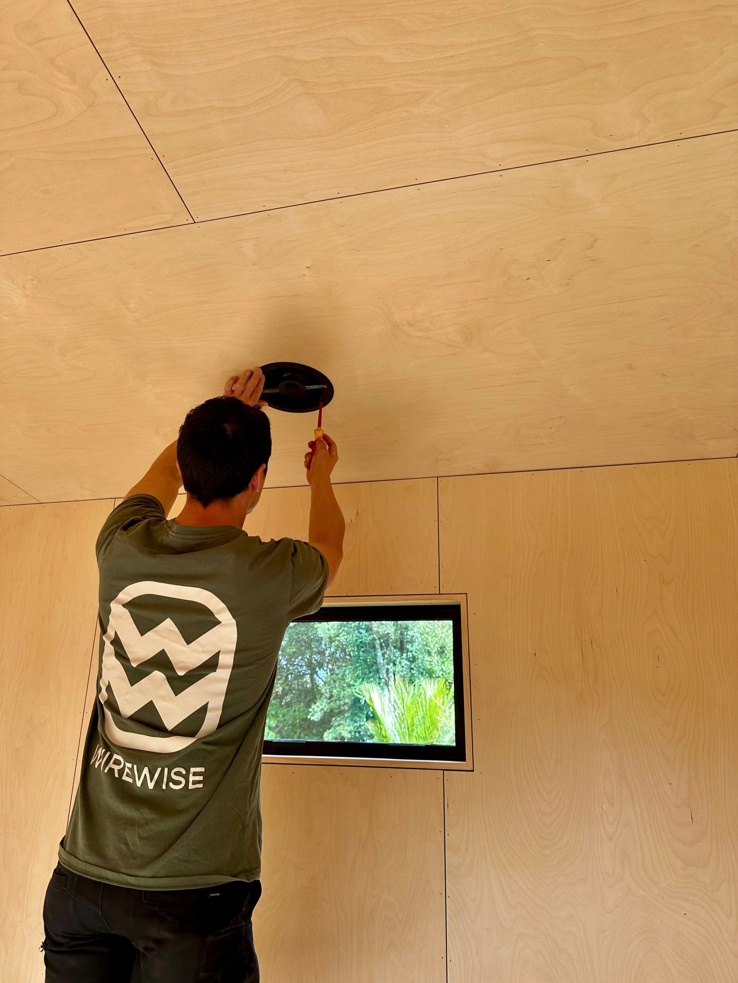 A person installing or repairing a ceiling light fixture in a room with wooden walls and a window showing greenery outside.