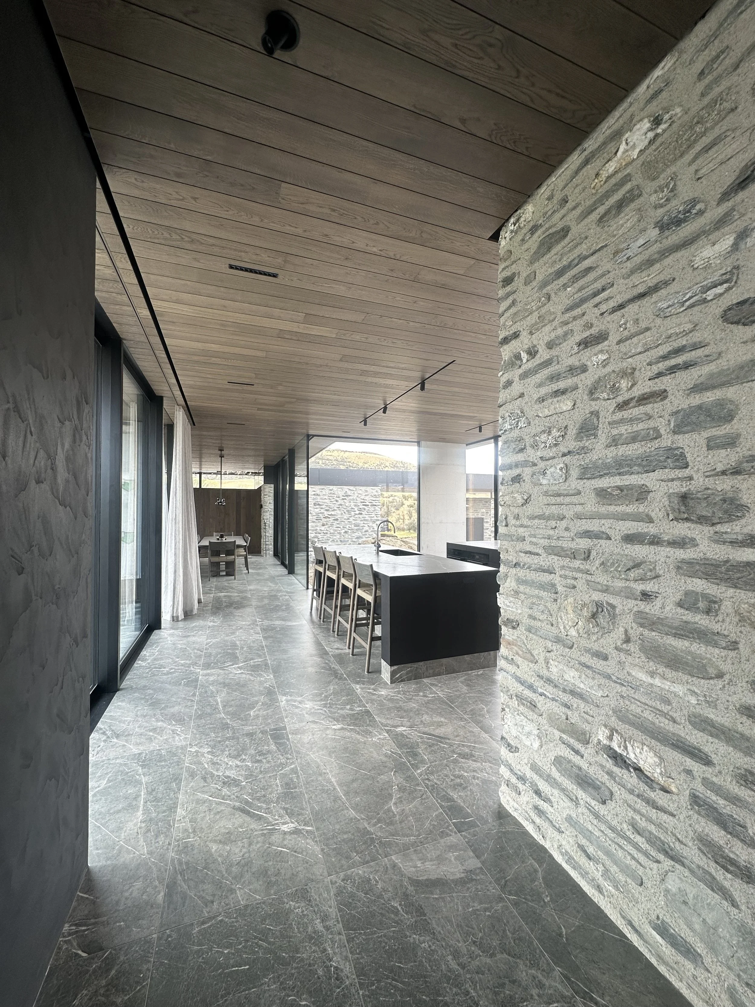 Modern interior with stone wall, wooden ceiling, marble floors, black kitchen island, and dining area with chairs and large windows.
