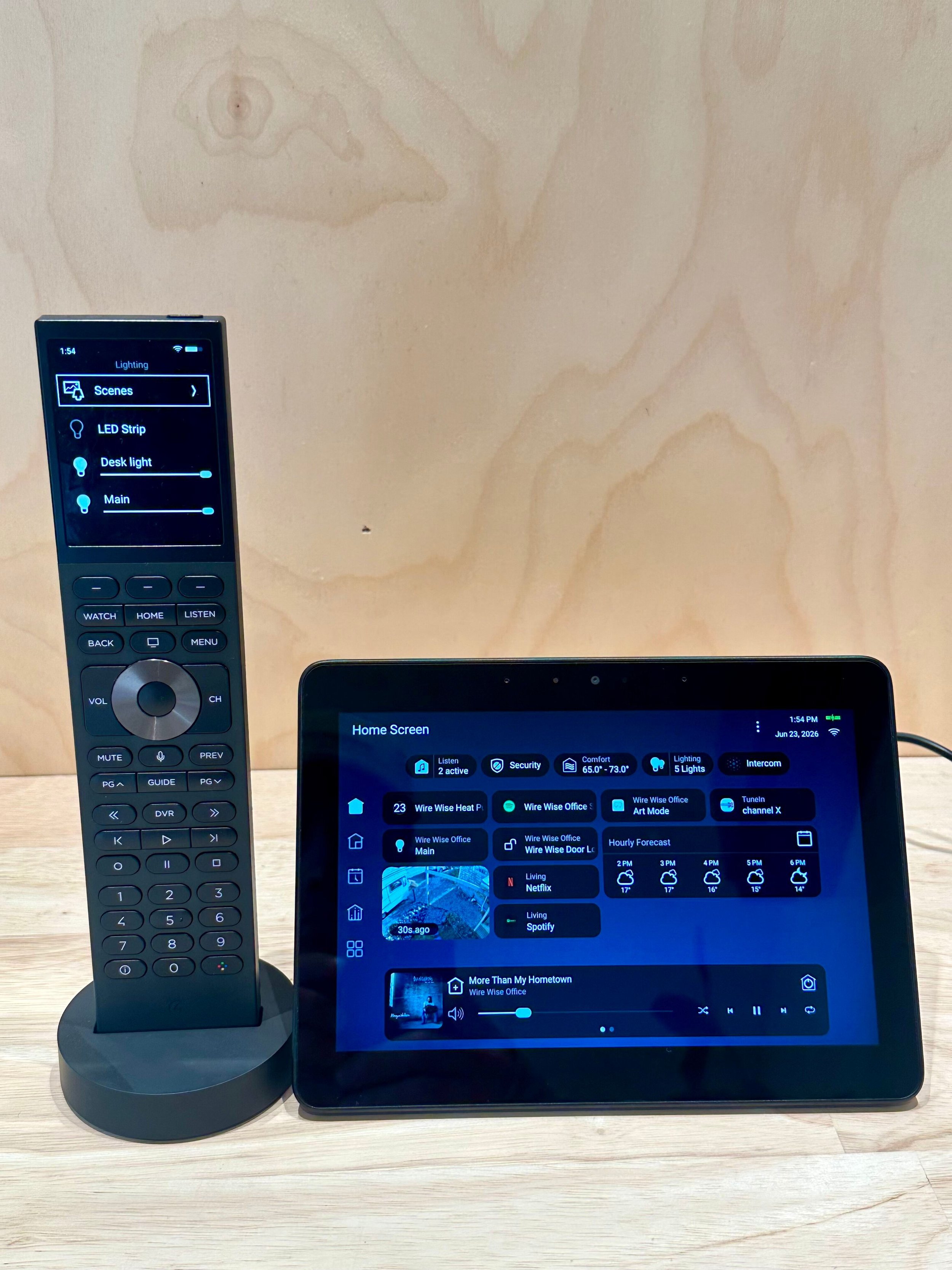 A remote control and a digital control panel on a wooden surface. The remote control is standing on a round base and displaying a menu screen with options like 'Scenes', 'LED Strip', and 'Desk light'. The digital panel shows a home screen with various icons and information, including weather, lighting, and media apps.