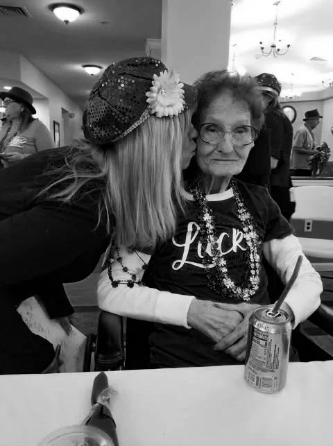 A young woman wearing a decorative headband and flower gives a kiss on the cheek to an elderly woman wearing glasses and a shirt with the word 'Lucky'. They are sitting at a table with a soda can and other items, in a lively indoor setting with other