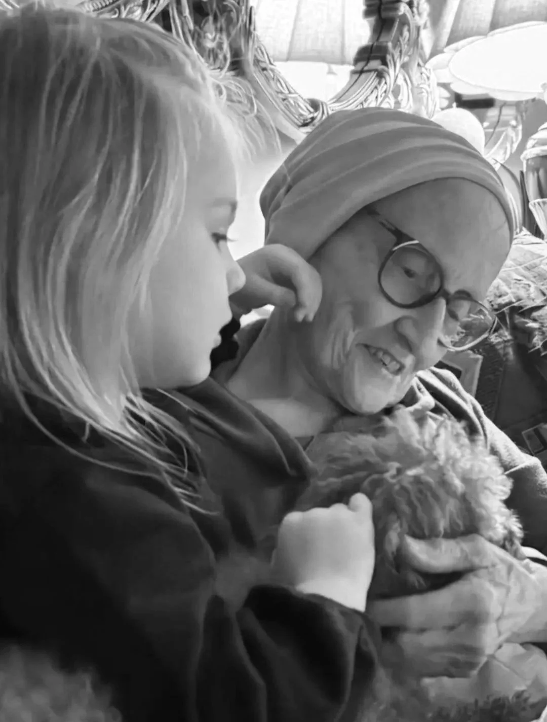 A young girl with long hair gently touching an elderly woman with glasses and a headscarf, both smiling. The elderly woman is holding a small dog. The scene is in a cozy indoor setting.
