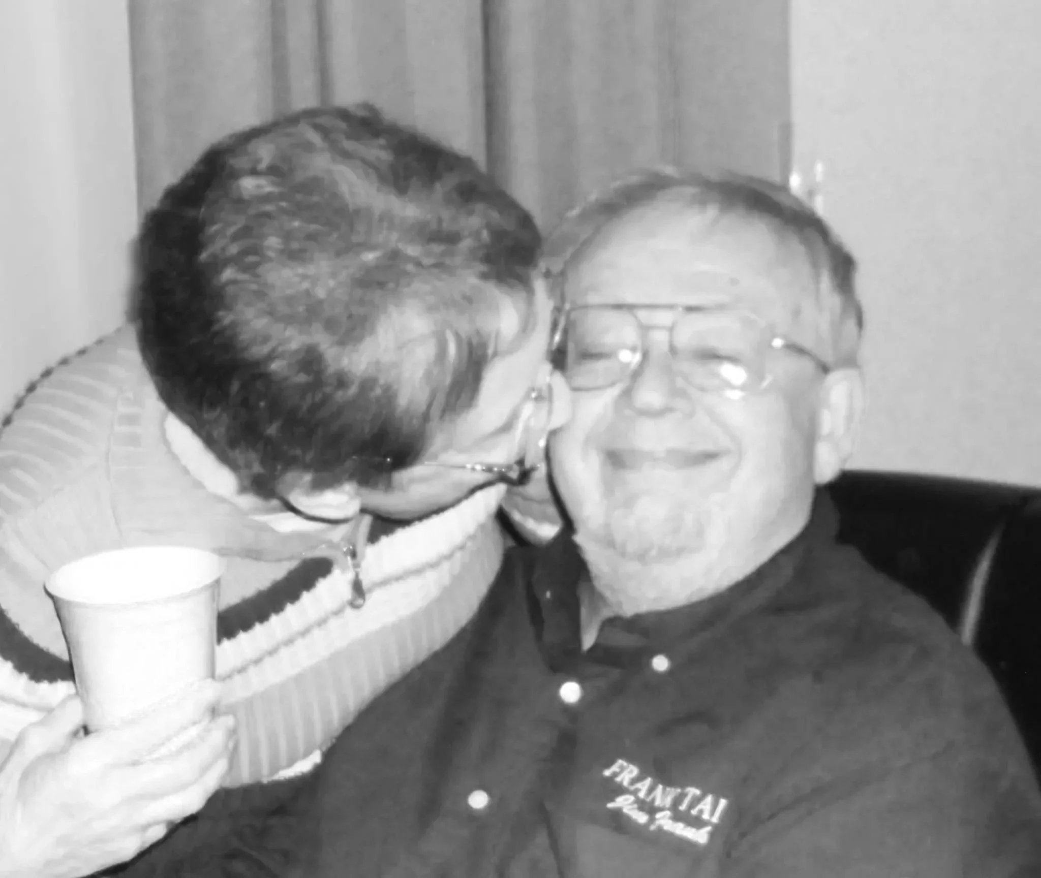 Two men smiling and sharing a moment of affection, one with glasses and the other with dark hair, in a black and white photo. One man is holding a cup.