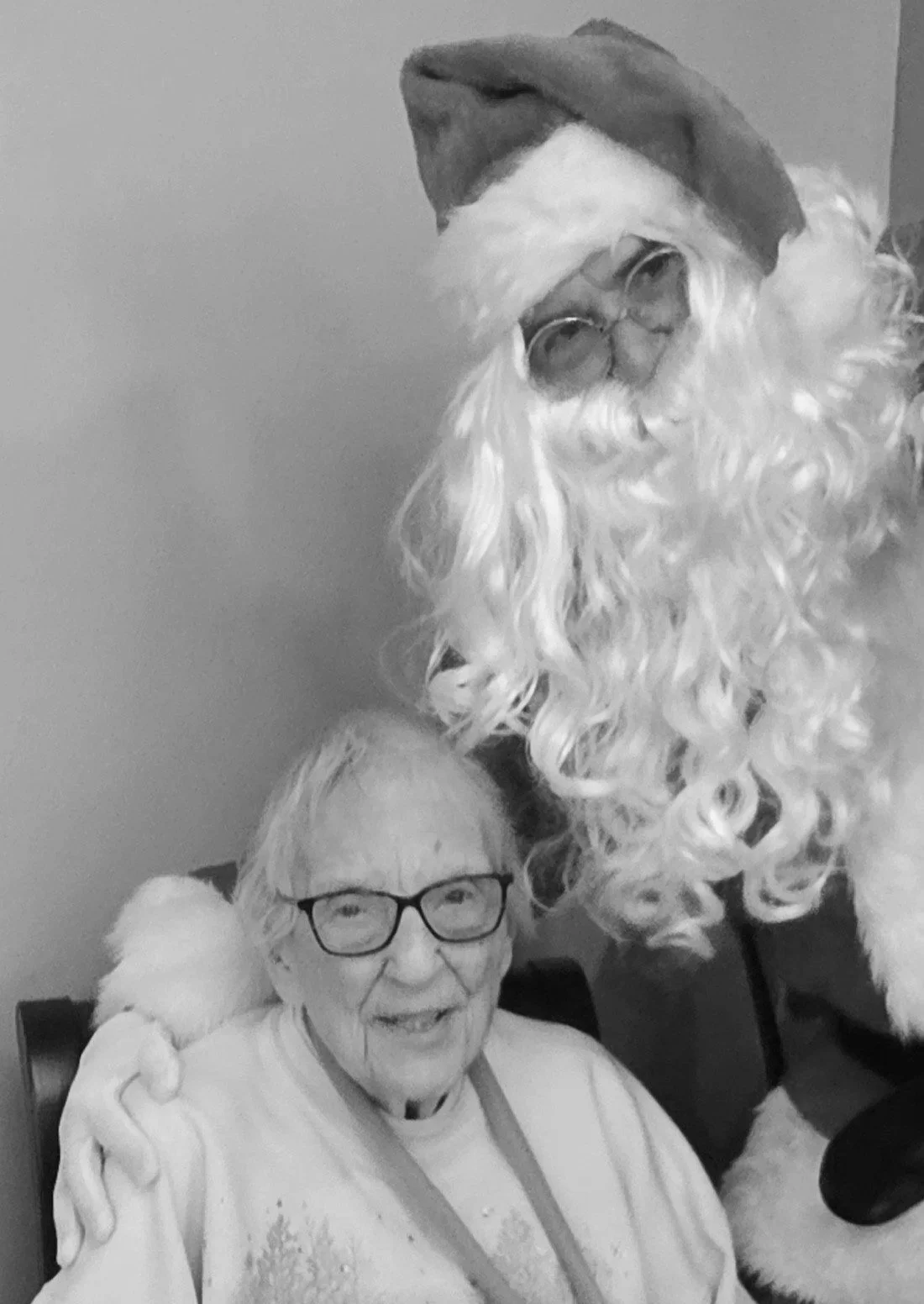 A woman with glasses smiling while sitting with a man dressed as Santa Claus, who is leaning towards her, in a black and white photo.