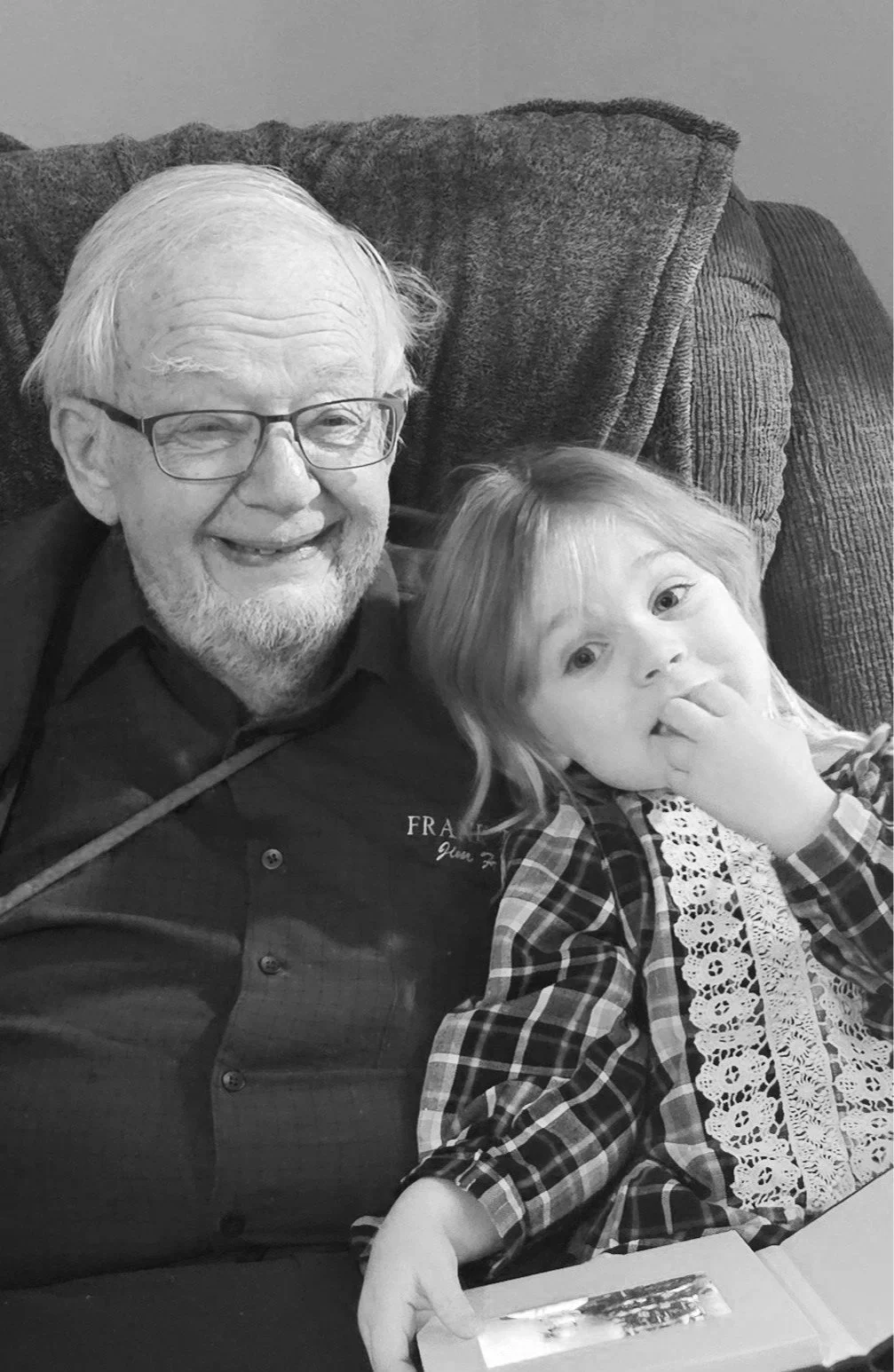 A black and white photo of an elderly man with glasses and a beard smiling, sitting on a couch with a young girl with light-colored hair, wearing a plaid dress with lace, who is touching her face and holding a picture frame.