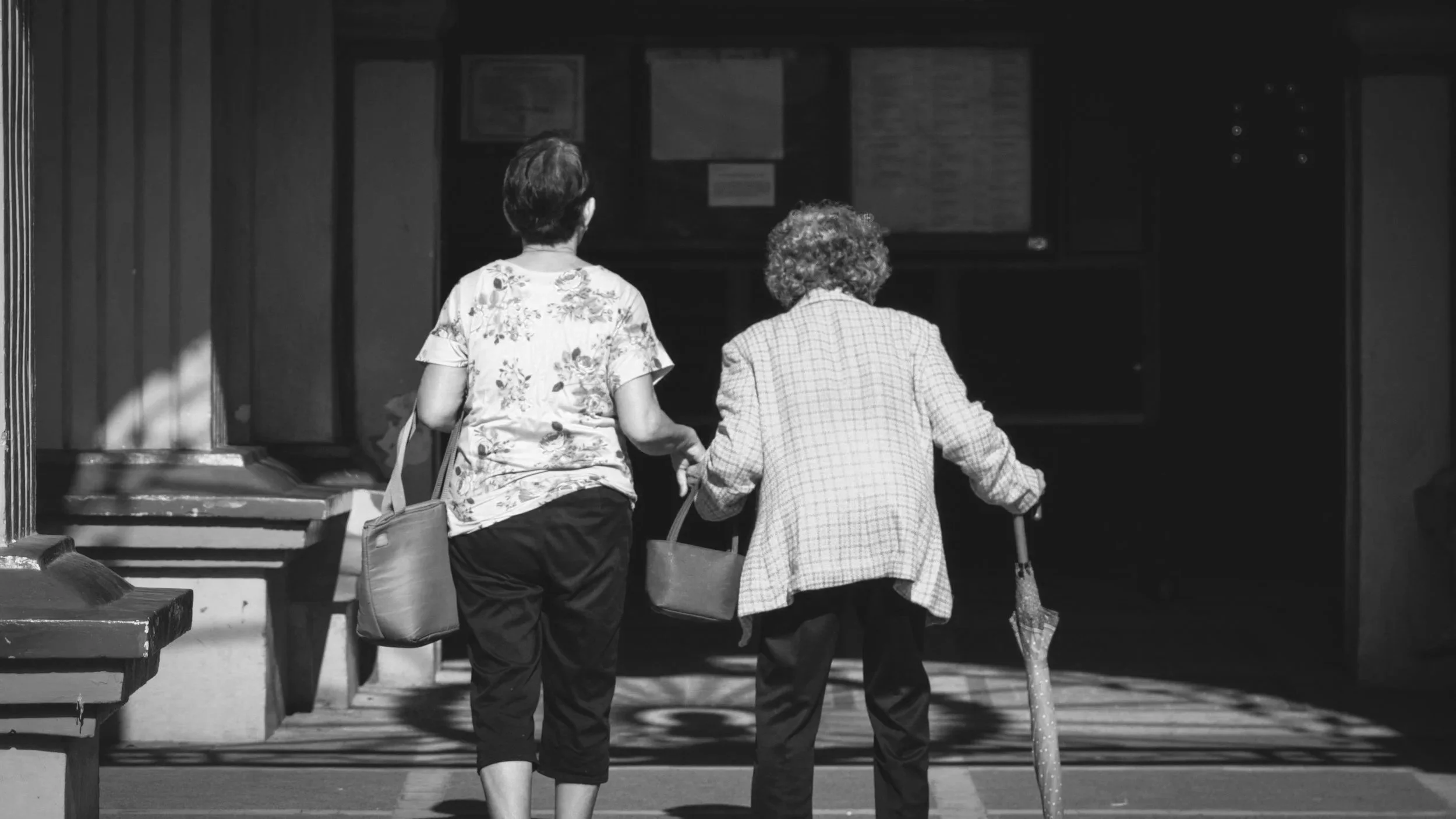 Two women seen from behind walking together, holding hands, with one using a cane, entering a building.