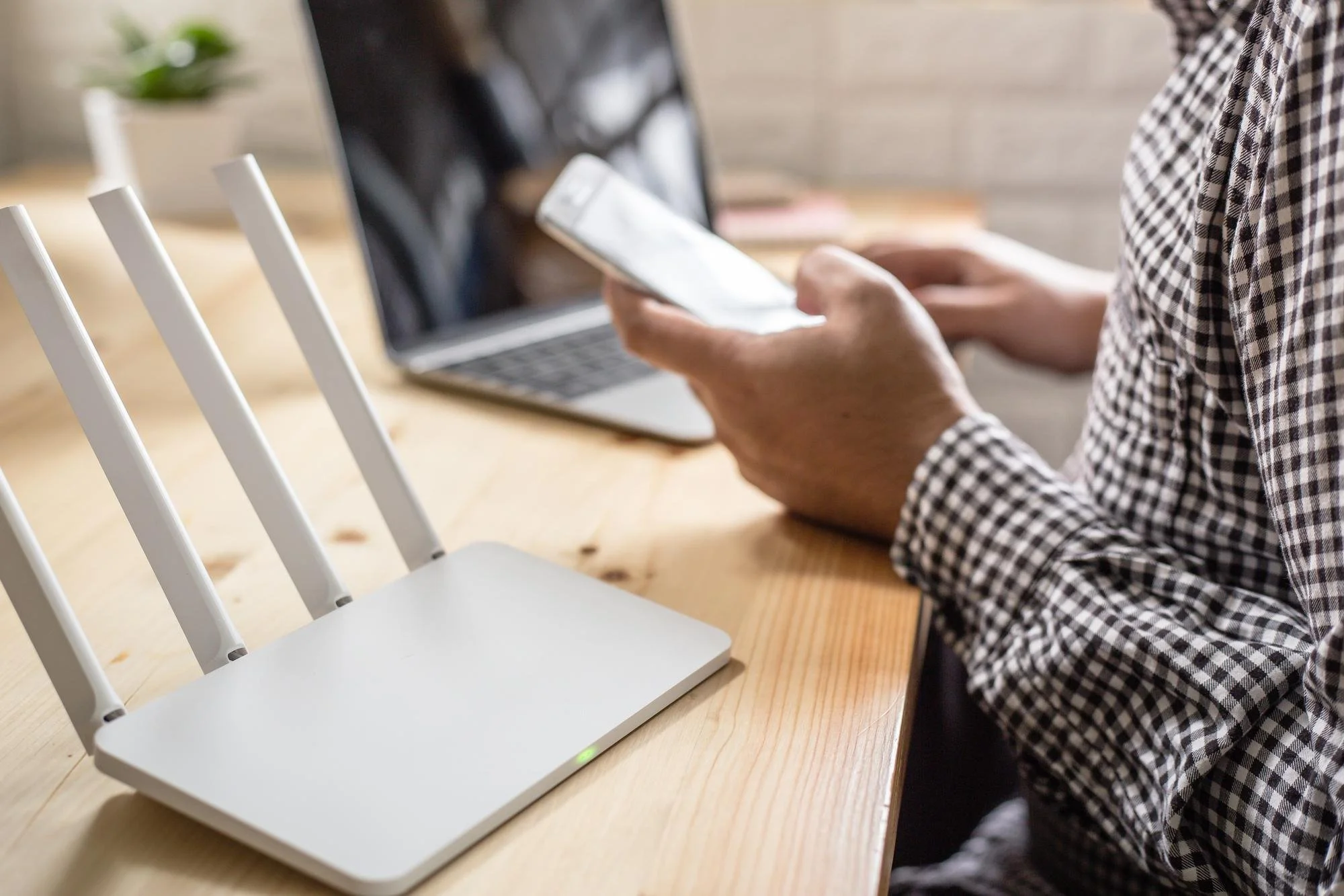 Person using a smartphone at a wooden table with a laptop and a wireless router.