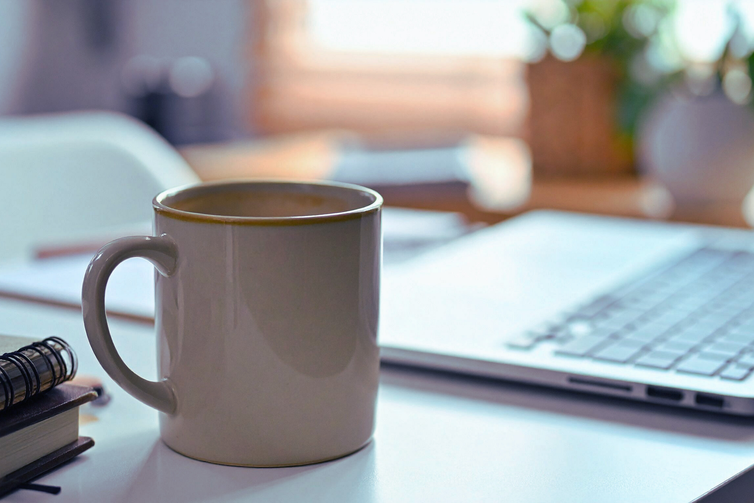 photograph of a coffee mug and notebooks in the foreground and a blurred laptop in the background