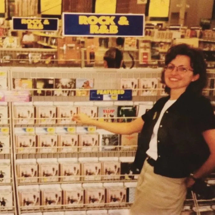 a photo of a young white woman with short dark hair and glasses standing in front of a CD display at a retail store