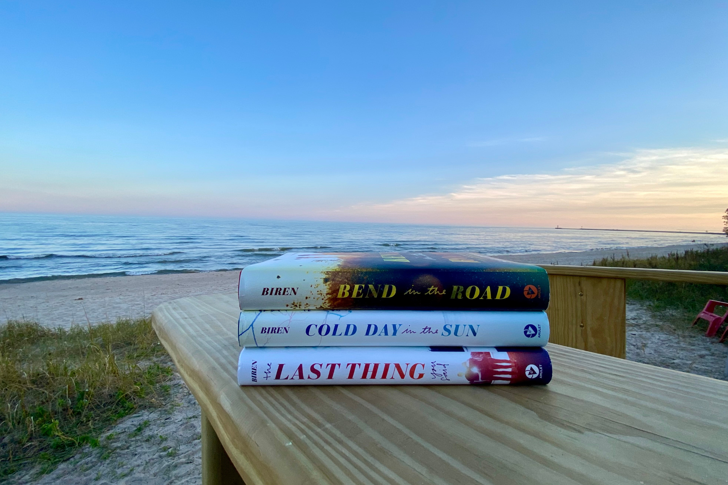 a stack of three hardcover novels by Sara Biren on the arm of a large wooden chair with a sandy beach and a lake at sunset in the background