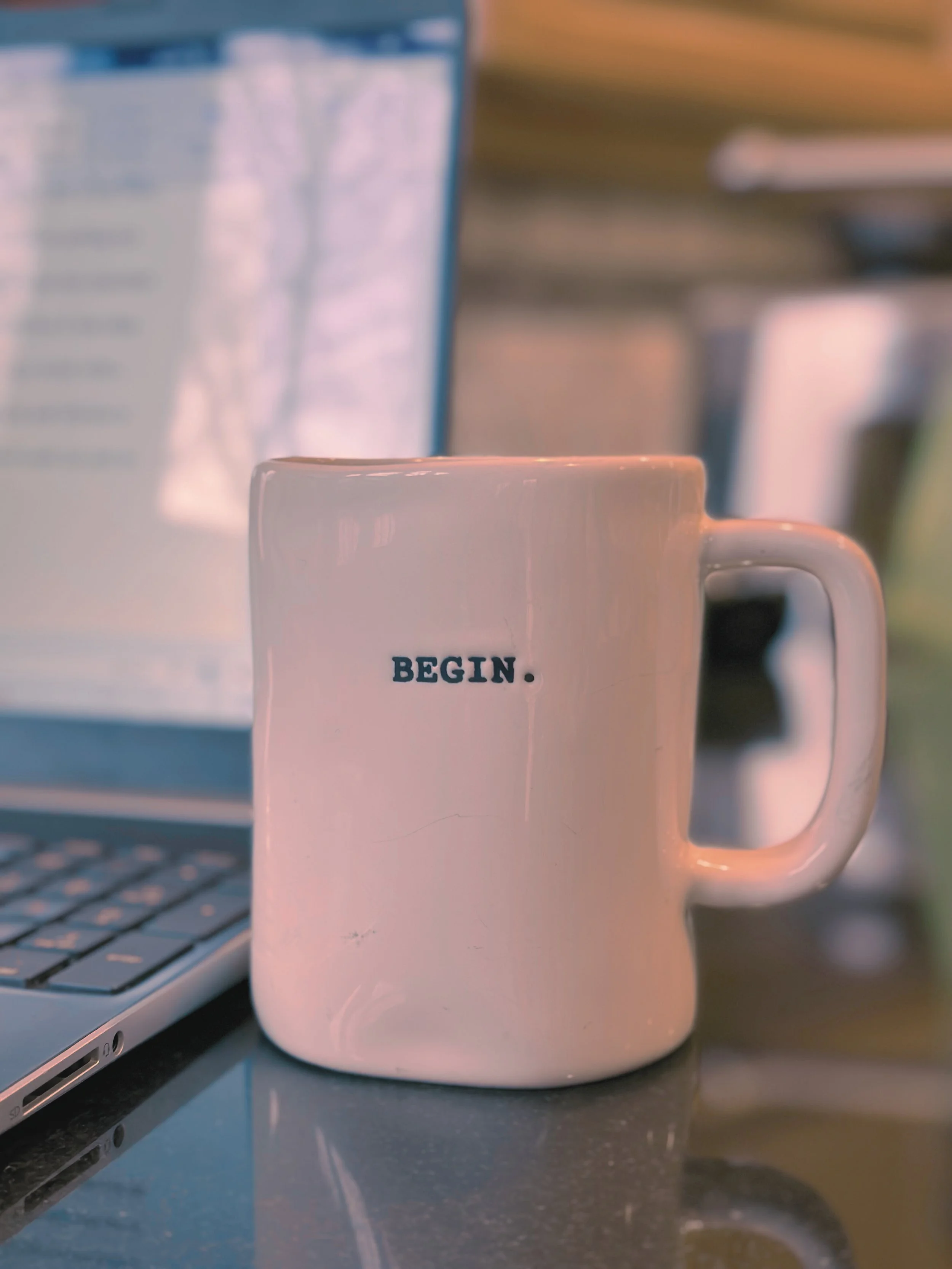 a photograph of a white mug with the word BEGIN. embossed in it in front of a laptop