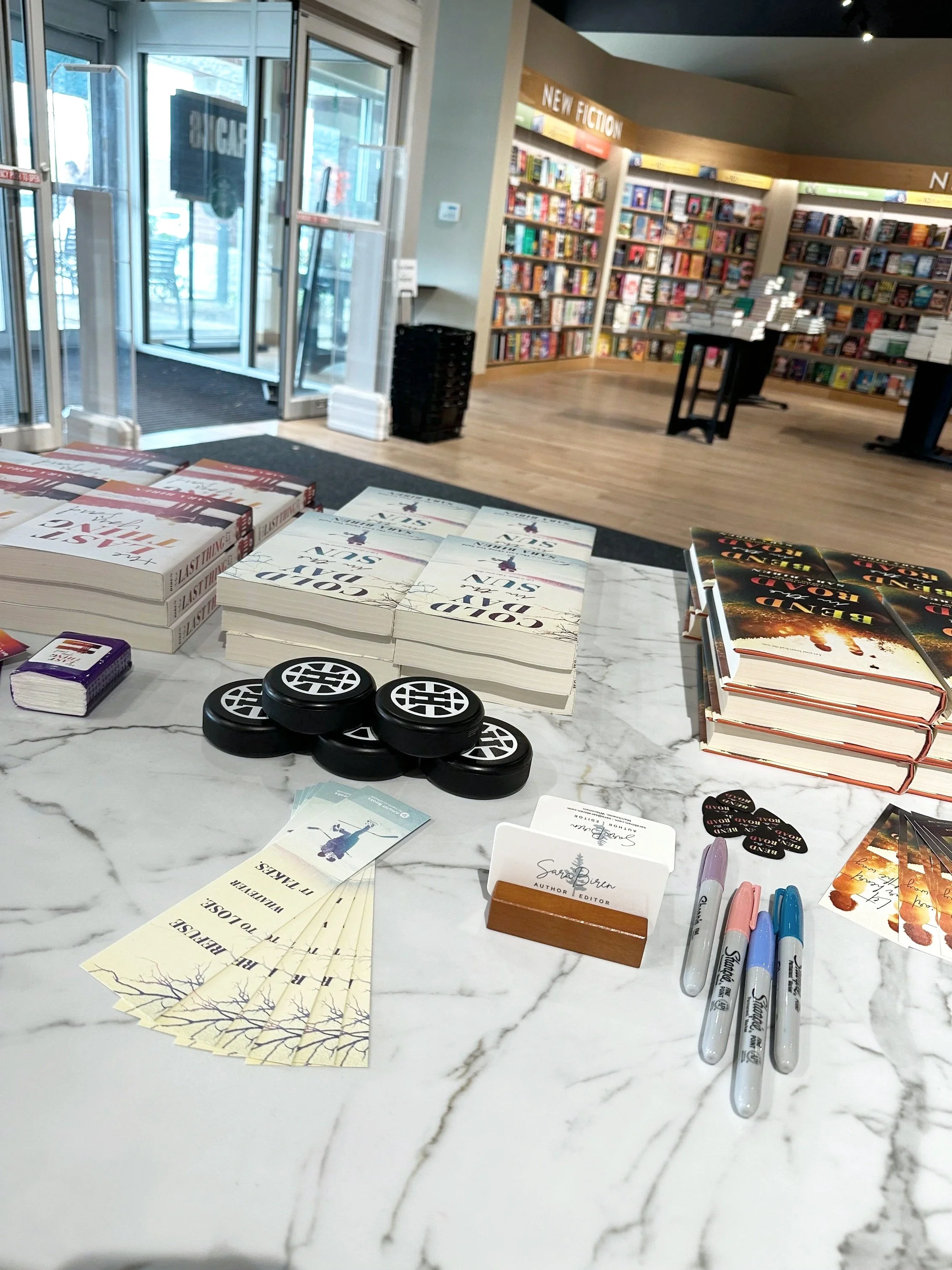 a photograph of stacks of books on a table along with promotional items at a bookstore with shelves and tables in the background
