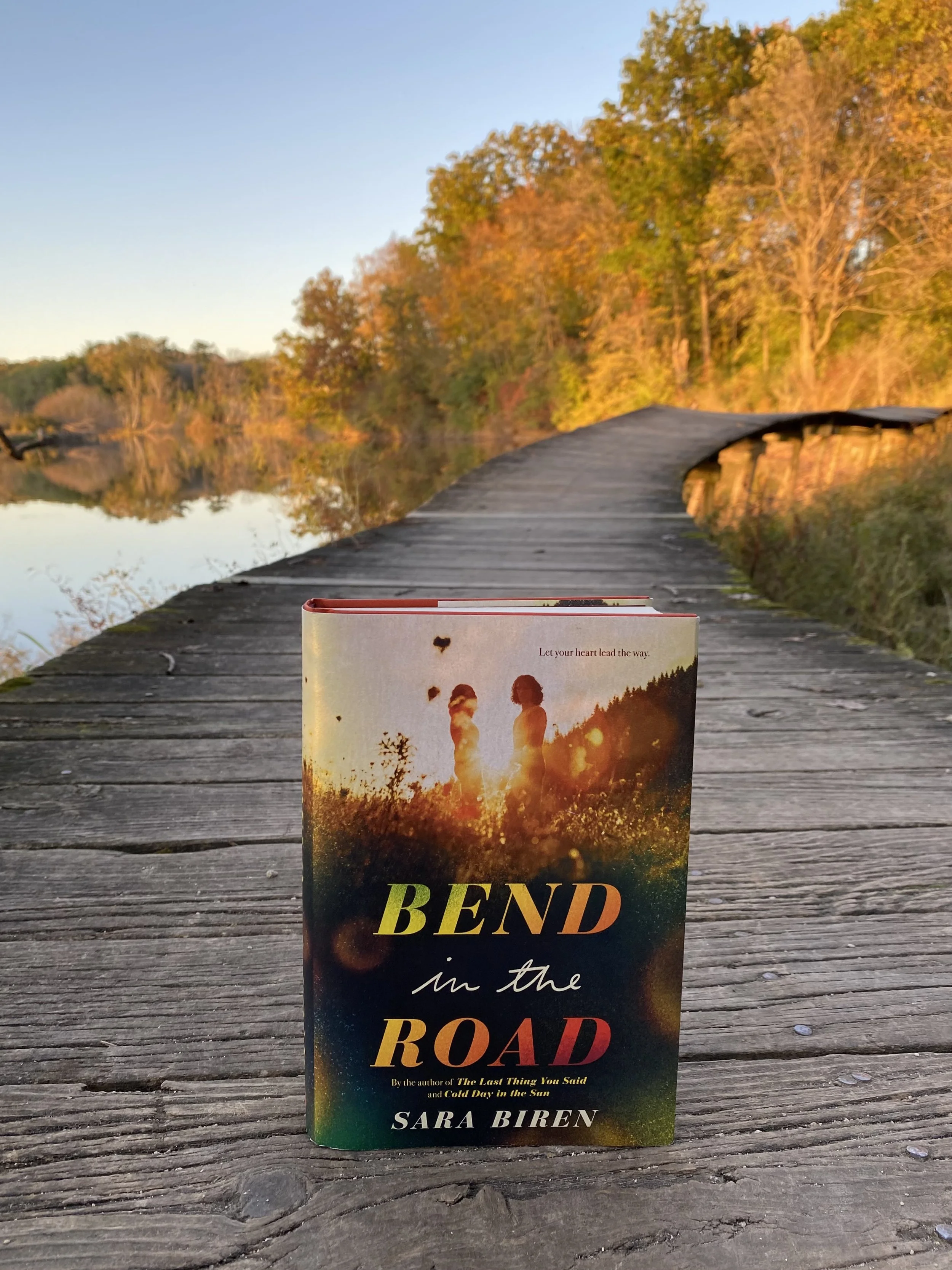 photograph of a hardcover book with the words Bend in the Road and Let your heart lead the way along with a female and male teenager standing in a sunlit meadow. The book sits on a wooden platform by a river and trees with orange leaves