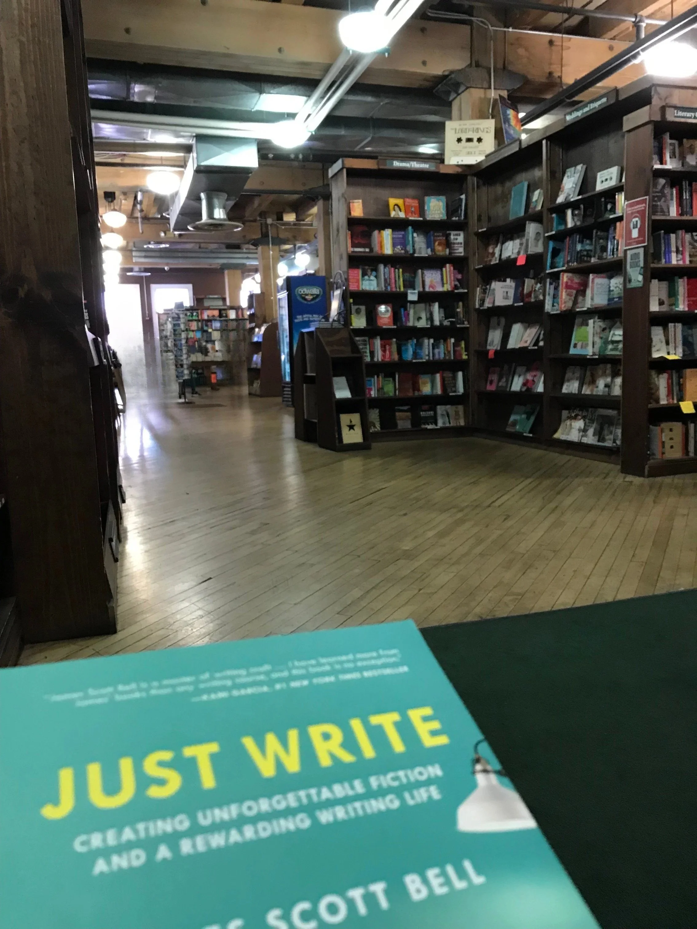 a photograph of a teal book cover with yellow letters that read JUST WRITE and a bookstore with shelves of books and wood floors in the background