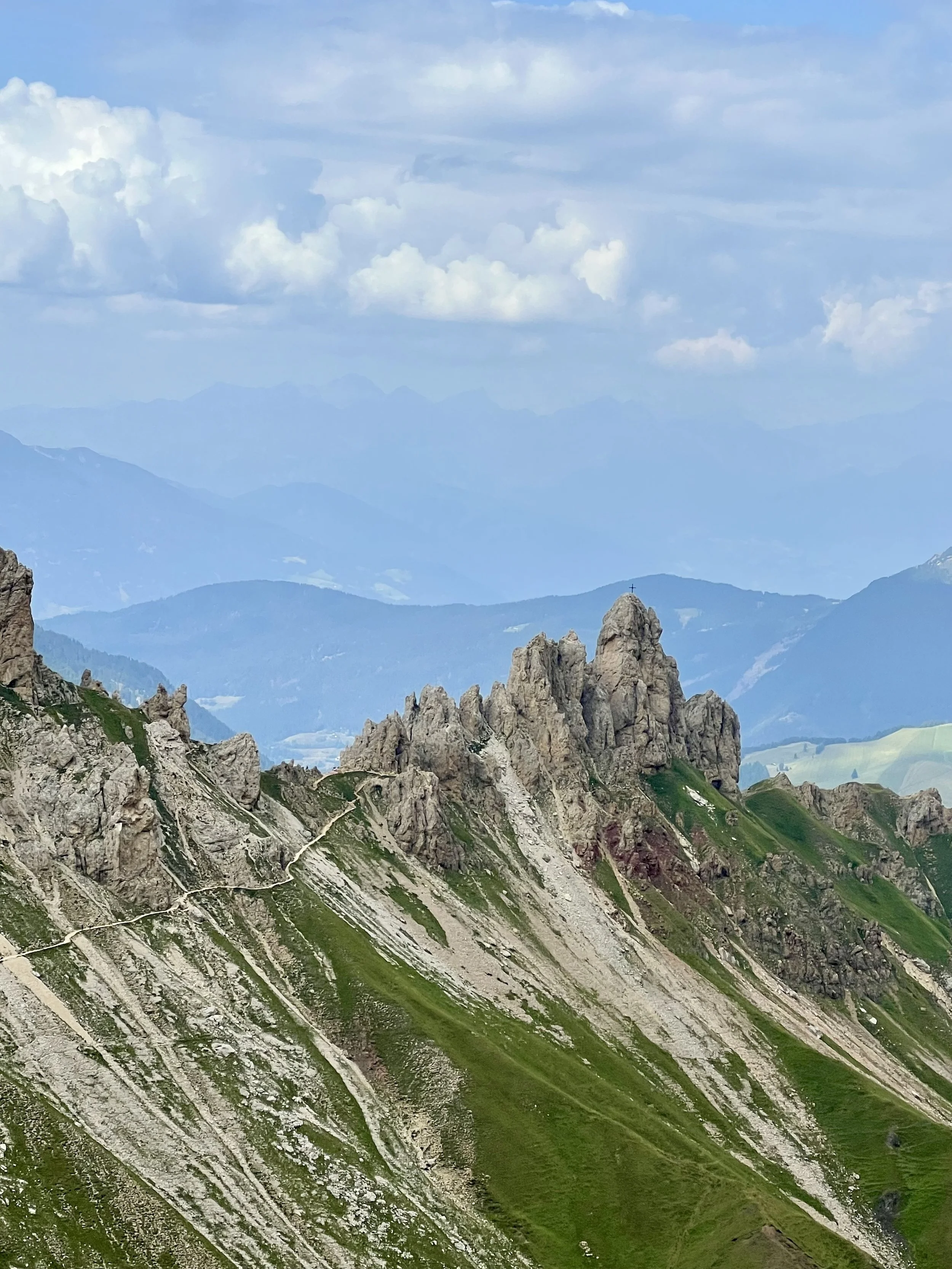 Mountain landscape with rocky peaks and green slopes, with distant blue mountain ranges under a cloudy sky.