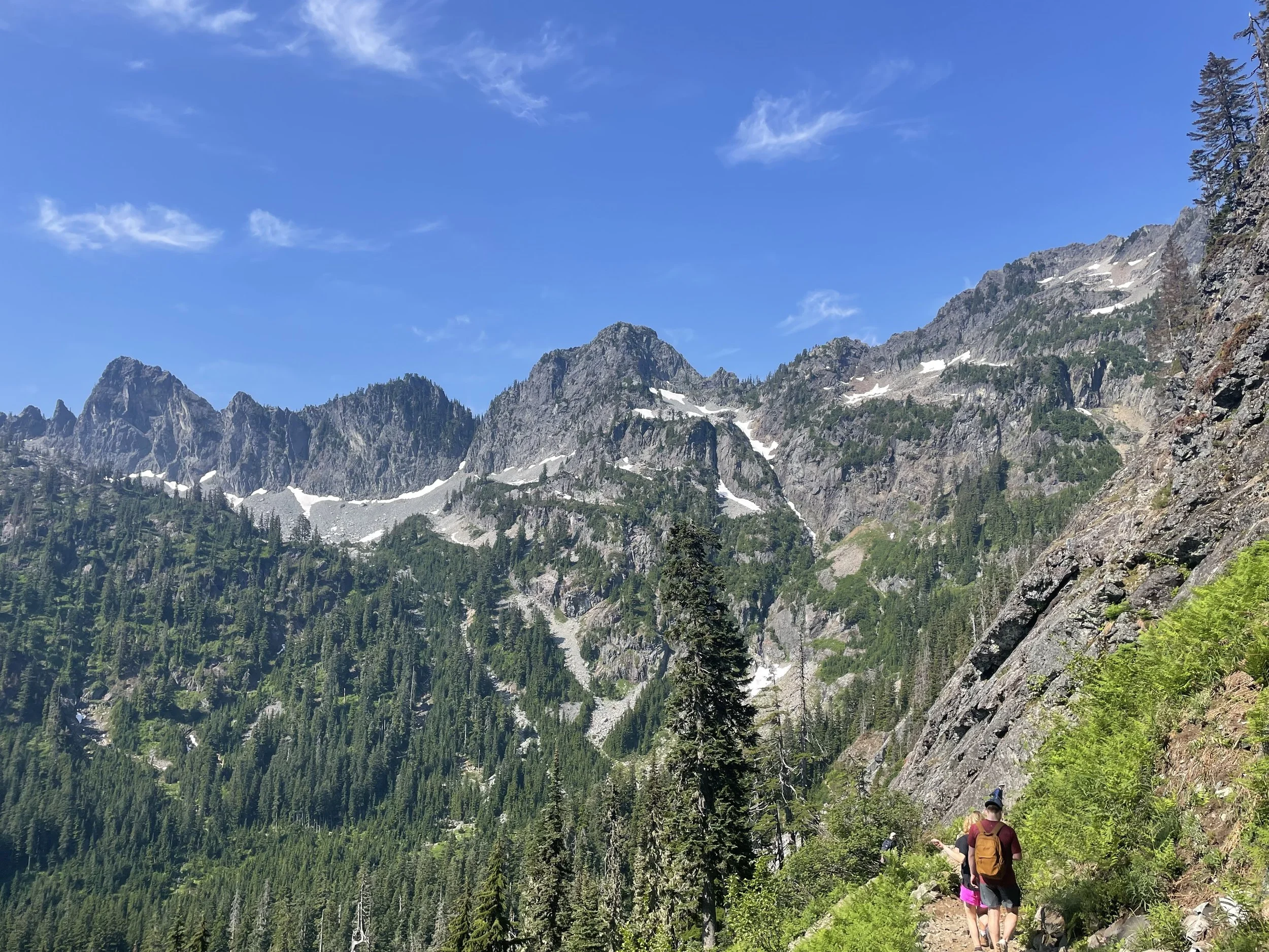 Hikers walking along a trail in a mountainous forested area with snow patches on rocky peaks and a clear blue sky.