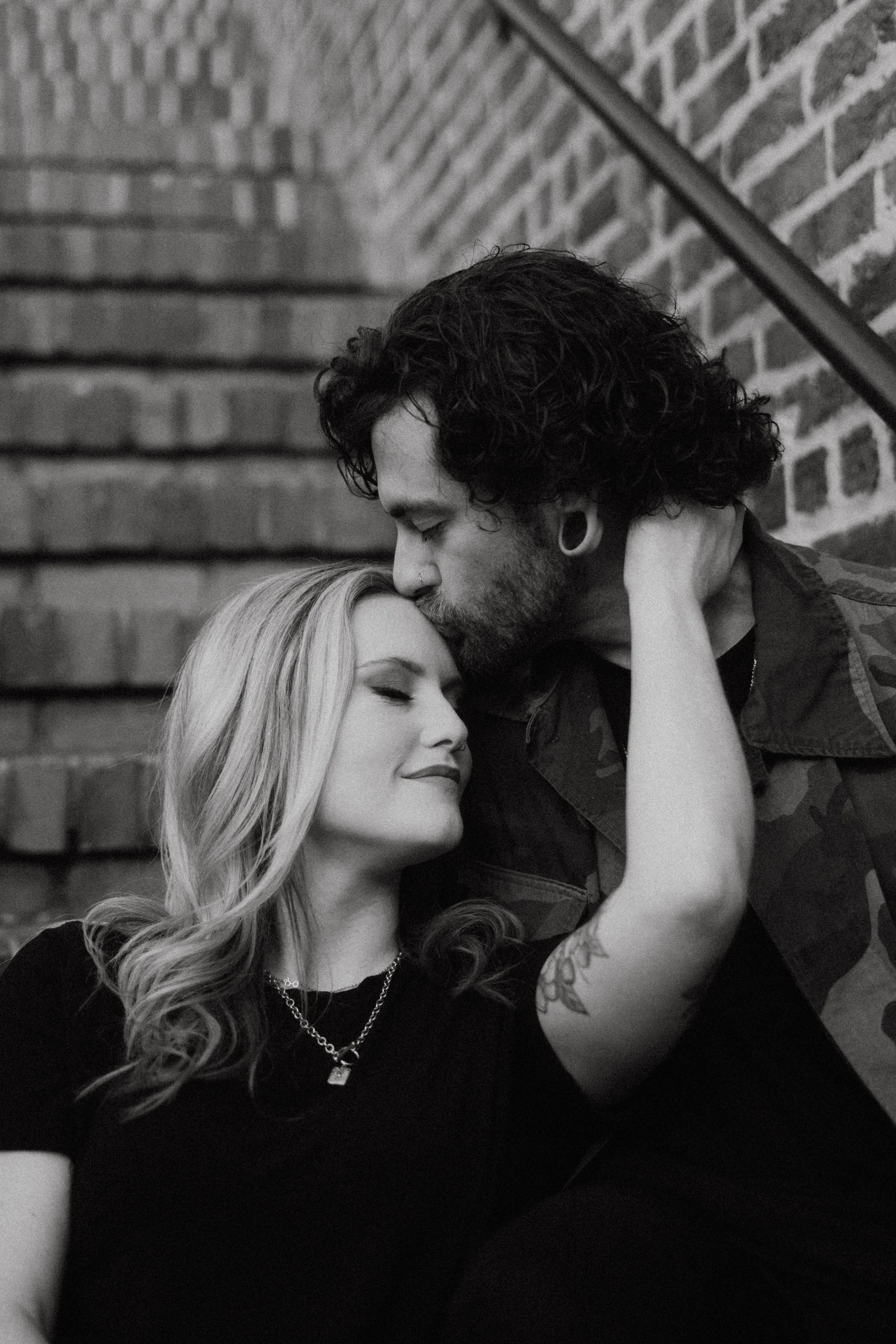 A black and white photo of a couple embracing, with the man kissing the woman's forehead as they both smile sweetly, sitting on brick stairs.