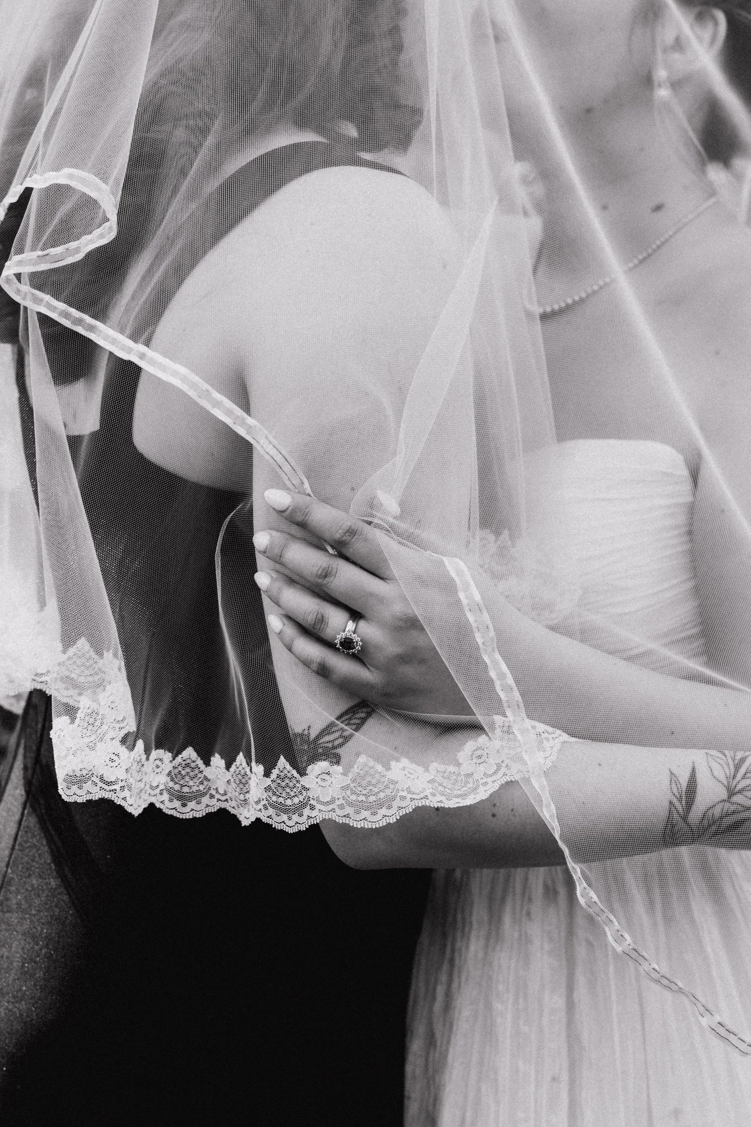 Close-up of a bride's arm and hand with an engagement ring, covered by a lace-edged veil, partially obscuring her face, in black and white.