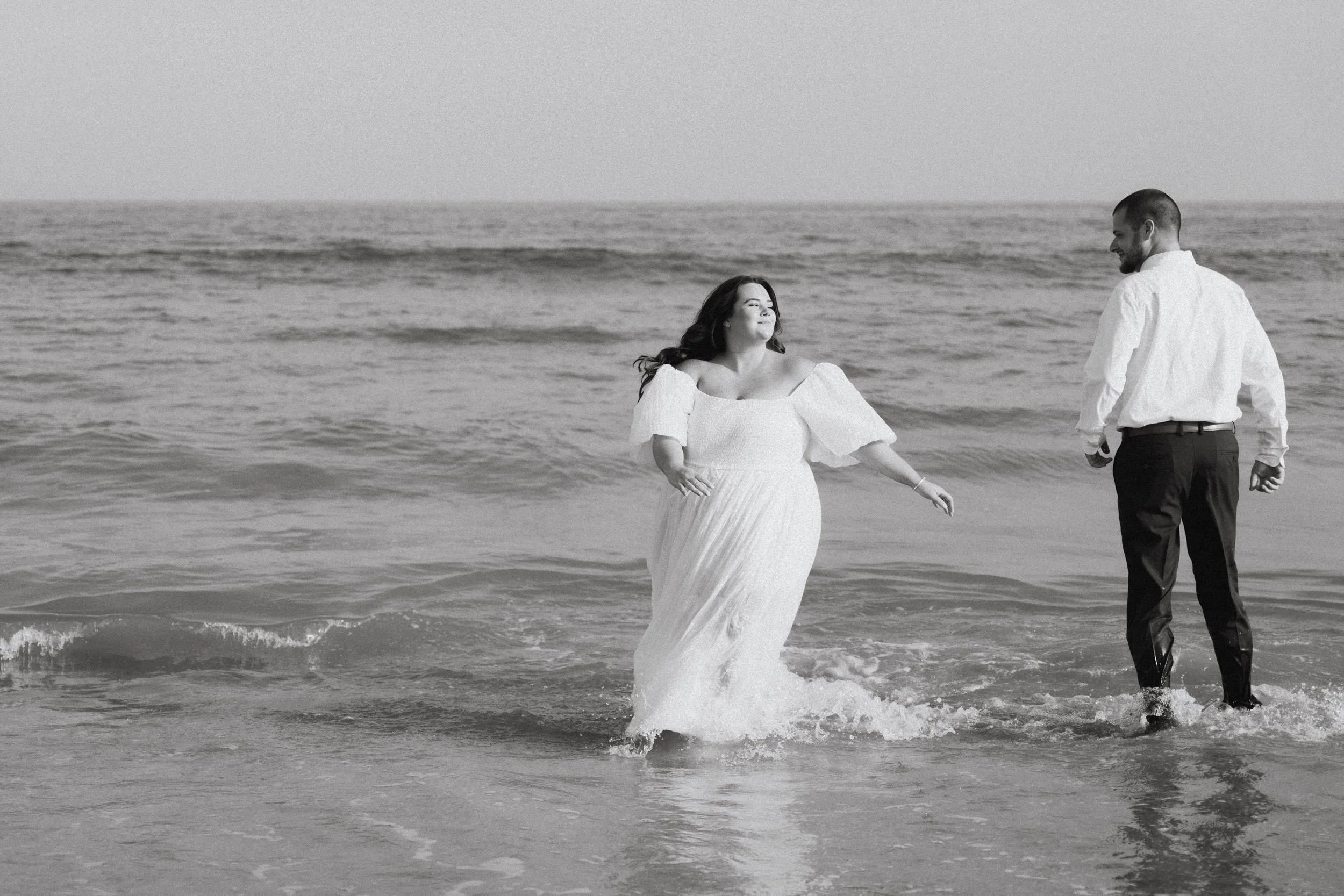 A woman in a white dress and a man in a white shirt with dark pants walk in the shallow ocean water at the beach. The woman is smiling and looking towards the man, who is looking down. The scene is in black and white.