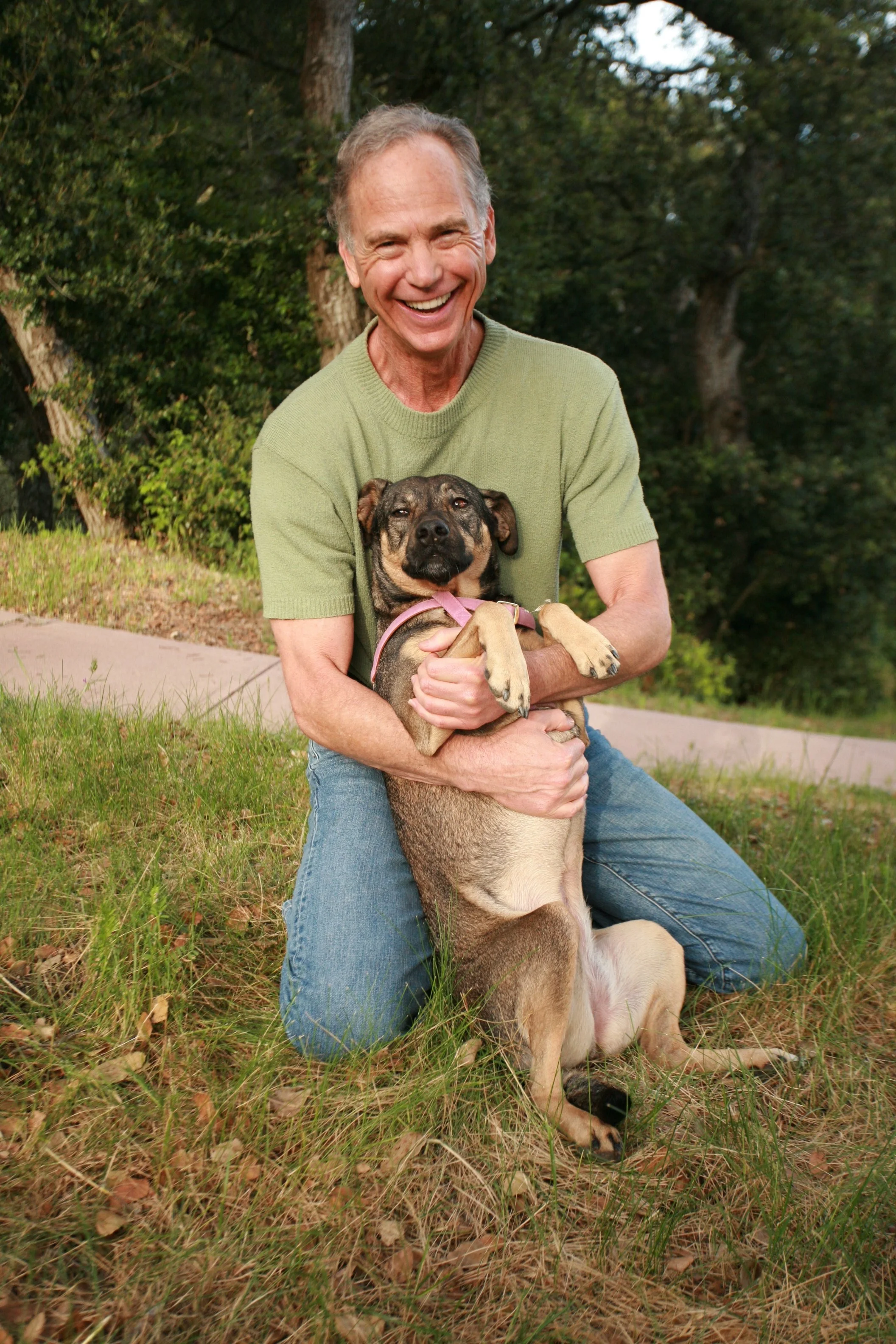 A man kneeling on the grass and smiling while hugging a large mixed breed dog with a brindle coat and tan markings in a park setting.
