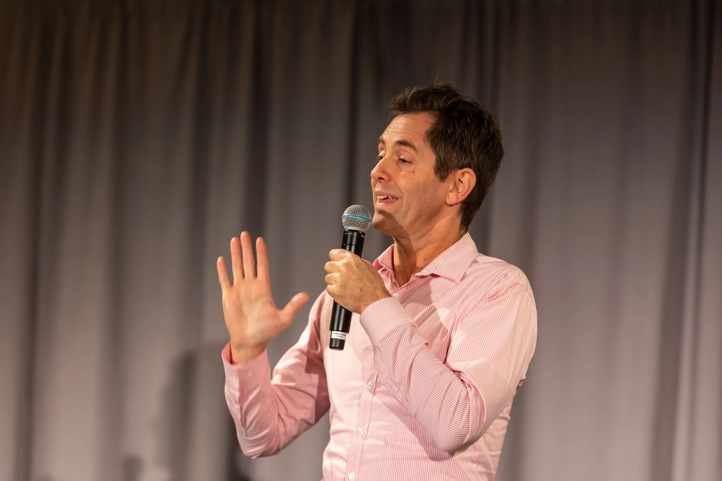A man with short dark hair, wearing a pink dress shirt, speaking into a microphone while gesturing with his right hand against a blurred curtain background.