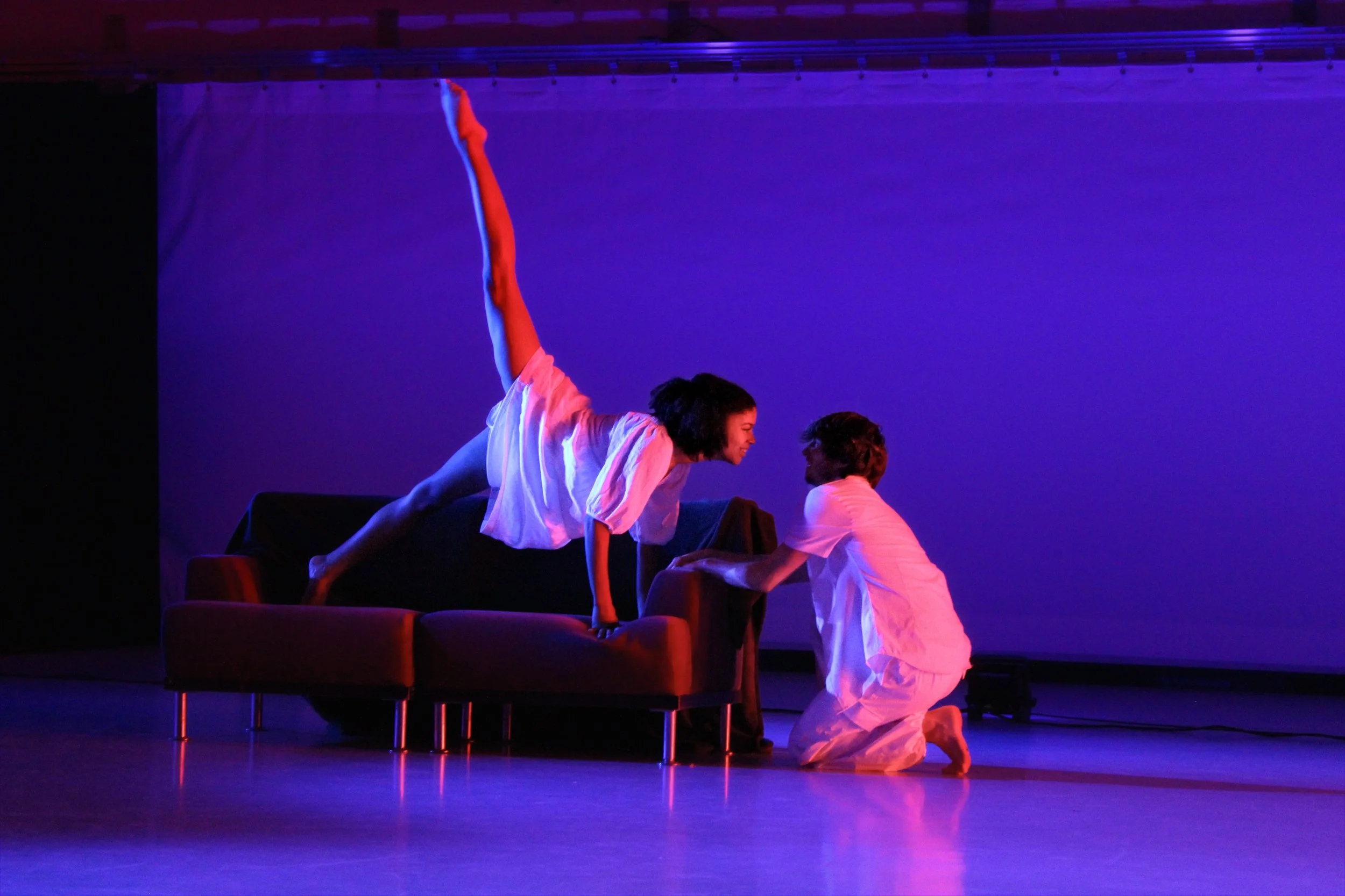 Performers in a stage act with colorful lighting, some crouching on the floor and one balancing on their hands and knees on a couch.