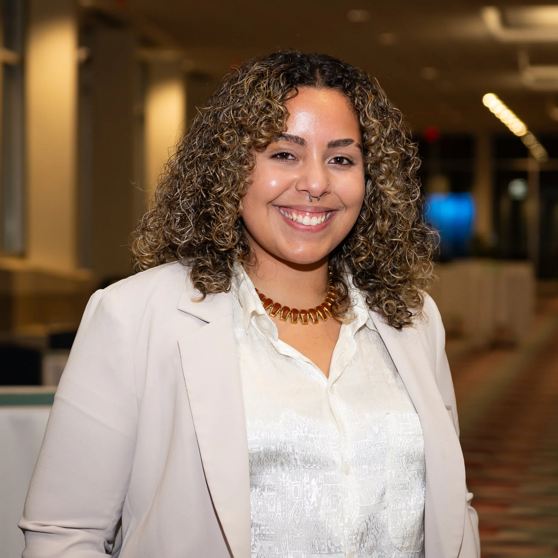A woman with curly hair and a septum piercing smiling, wearing a white blazer and a gold necklace, in a well-lit indoor space.